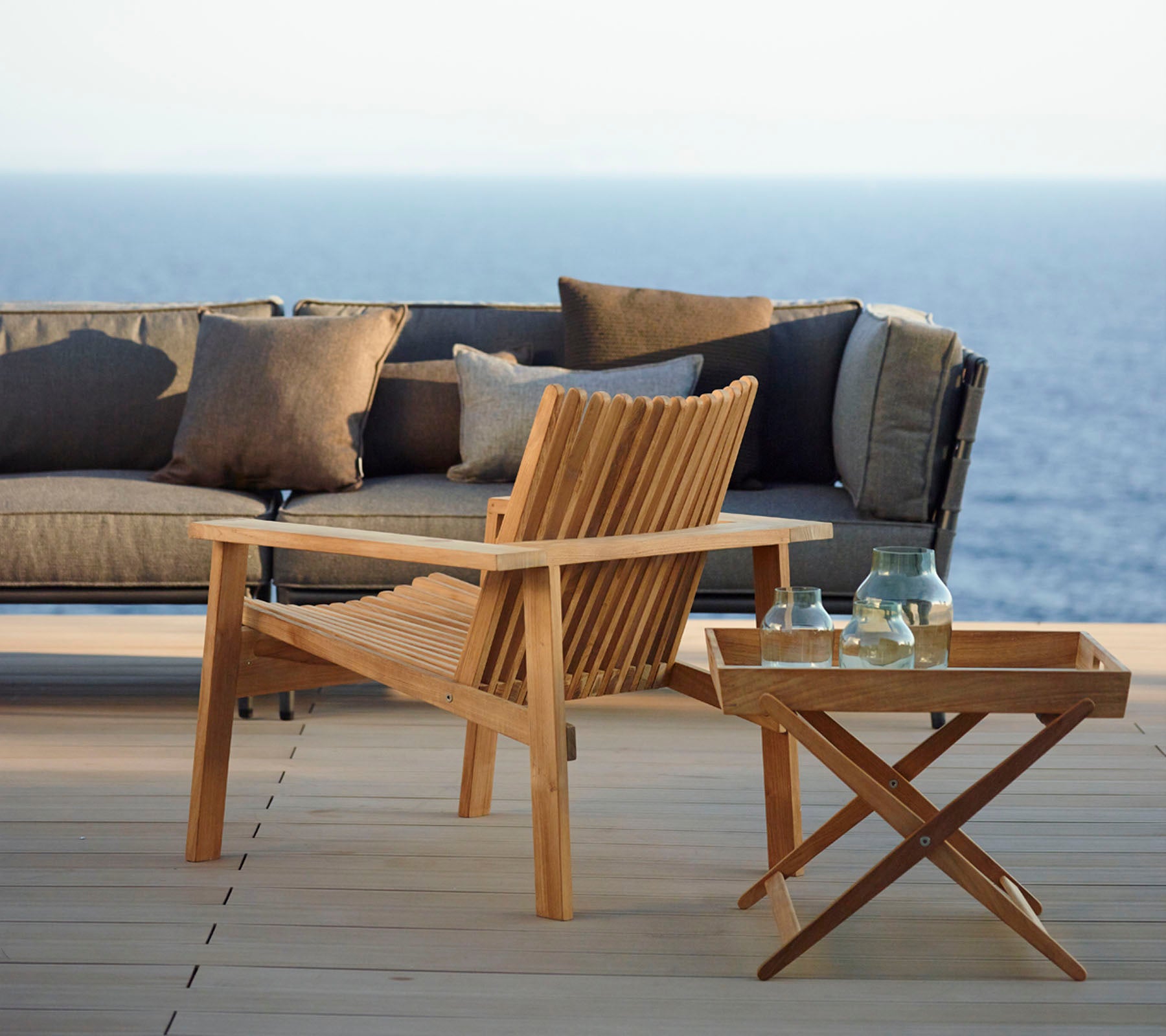 Chaise en bois élégante et table d'appoint avec des bouteilles, accompagnées d'un canapé moderne orné de coussins, le tout sur fond d'une mer paisible.