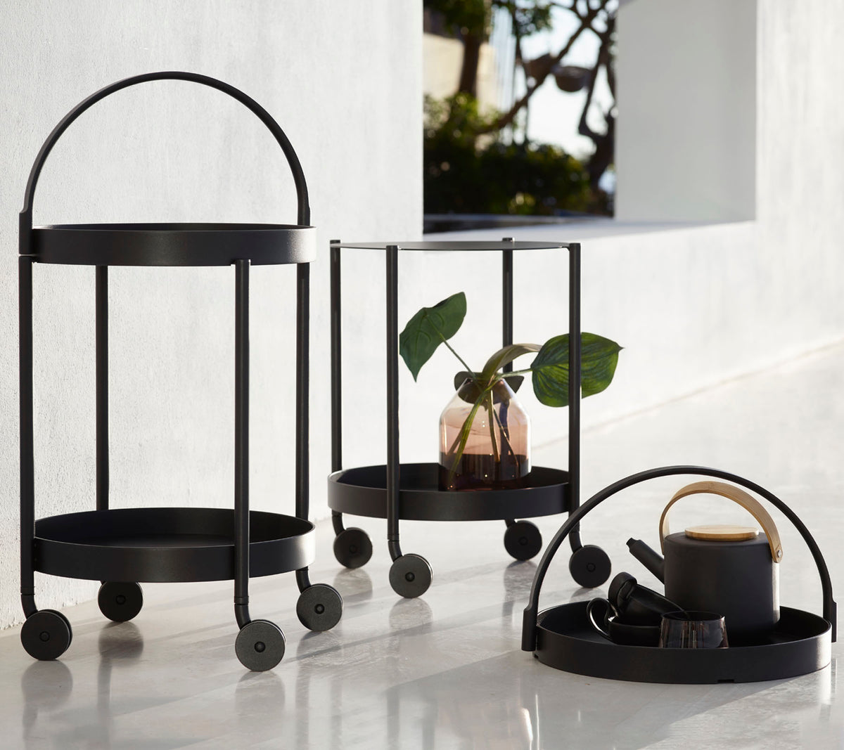 Trois chariots en métal noir avec des cadres circulaires et des roues, comprenant une plante, des objets décoratifs et une tasse, disposés pour l'affichage.