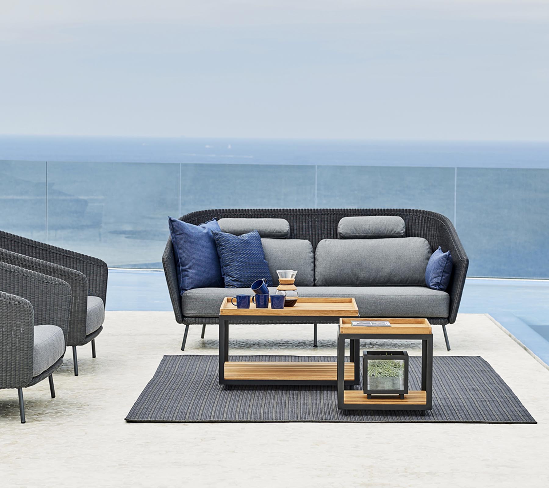 Agencement de sièges extérieur élégant avec un canapé gris et des coussins bleus, une table d'appoint en bois et un tapis assorti.