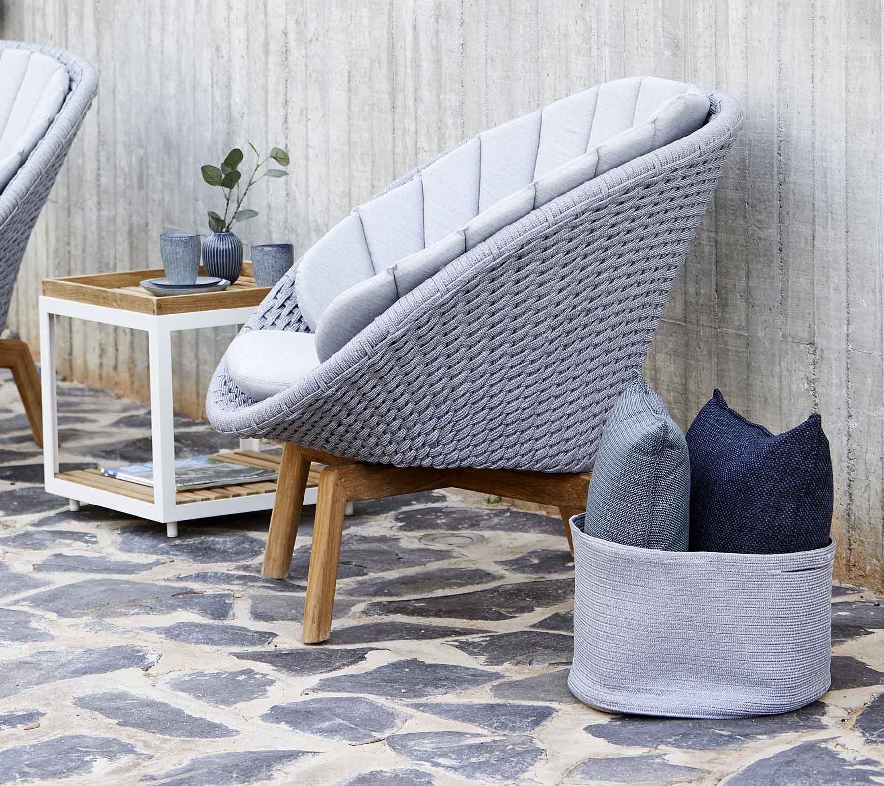 Fauteuil moderne gris avec un design tissé, accompagné de coussins et d'une petite table d'appoint en bois. Le sol en pierre améliore l'esthétique.