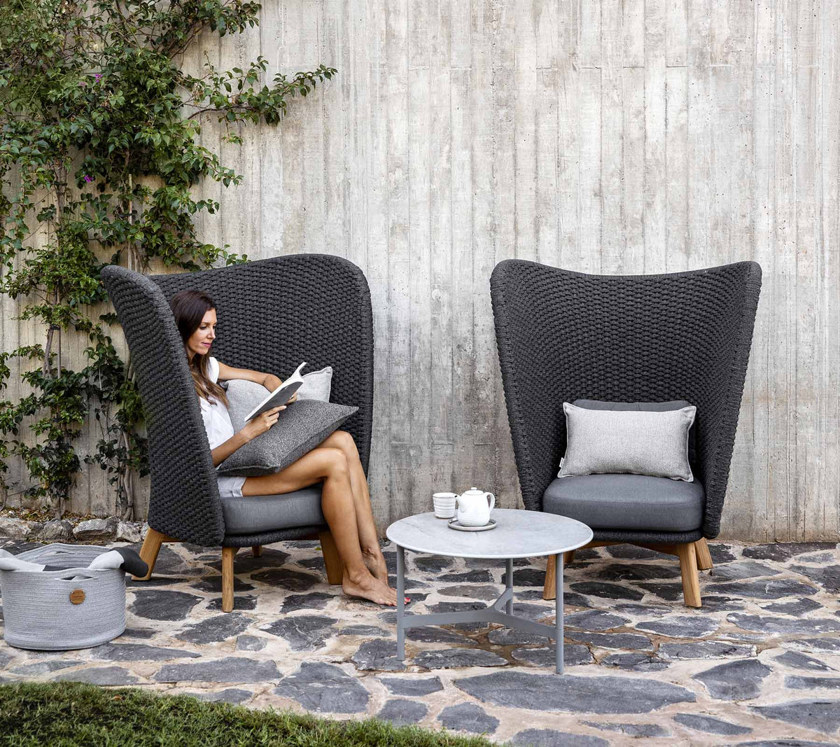Une femme lit confortablement assise dans une chaise en rotin élégante à côté d'une petite table, entourée de verdure et de pierres lisses.