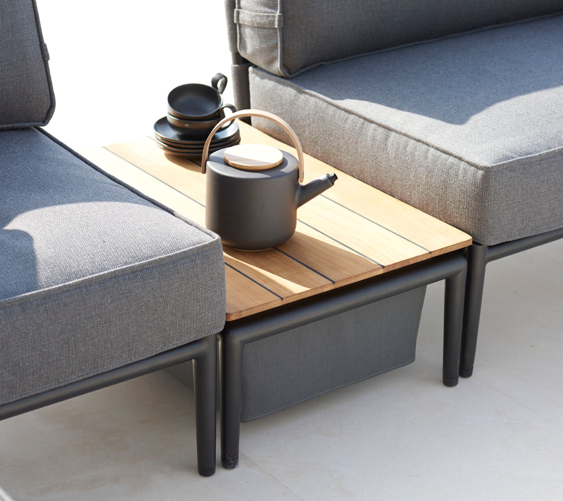 Deux canapés gris flanquent une table d'appoint en bois, surmontée d'une théière noire et de plusieurs tasses, créant un coin salon chaleureux.