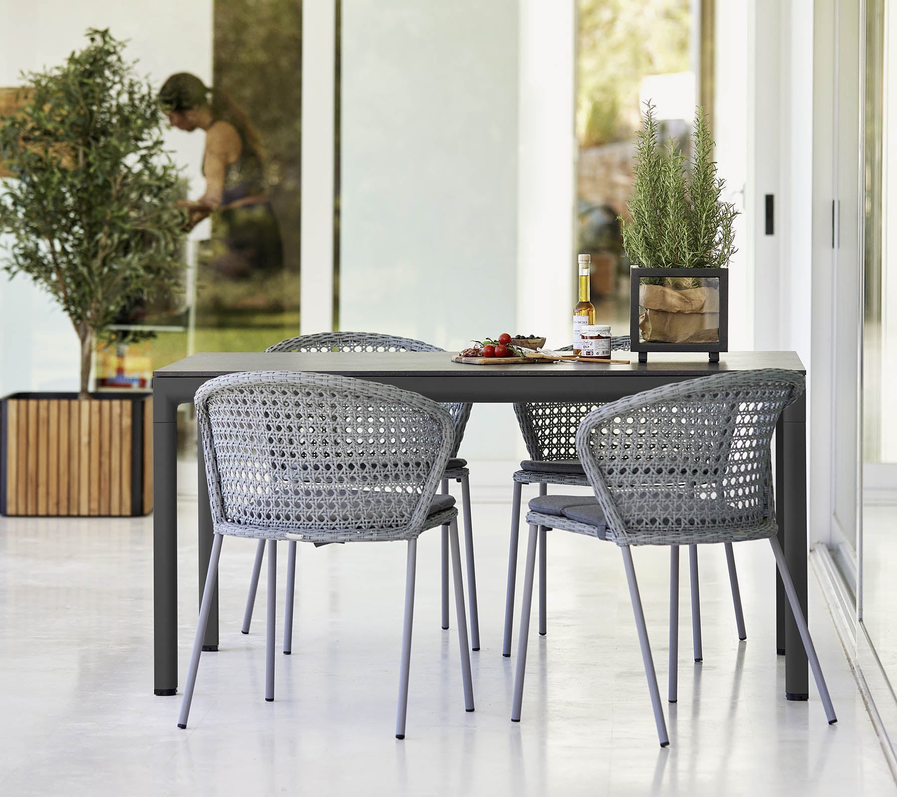 Ensemble de table moderne avec quatre chaises tressées grises, ornées de petites plantes et d'un centre de table décoratif. Ambiance lumineuse et accueillante.