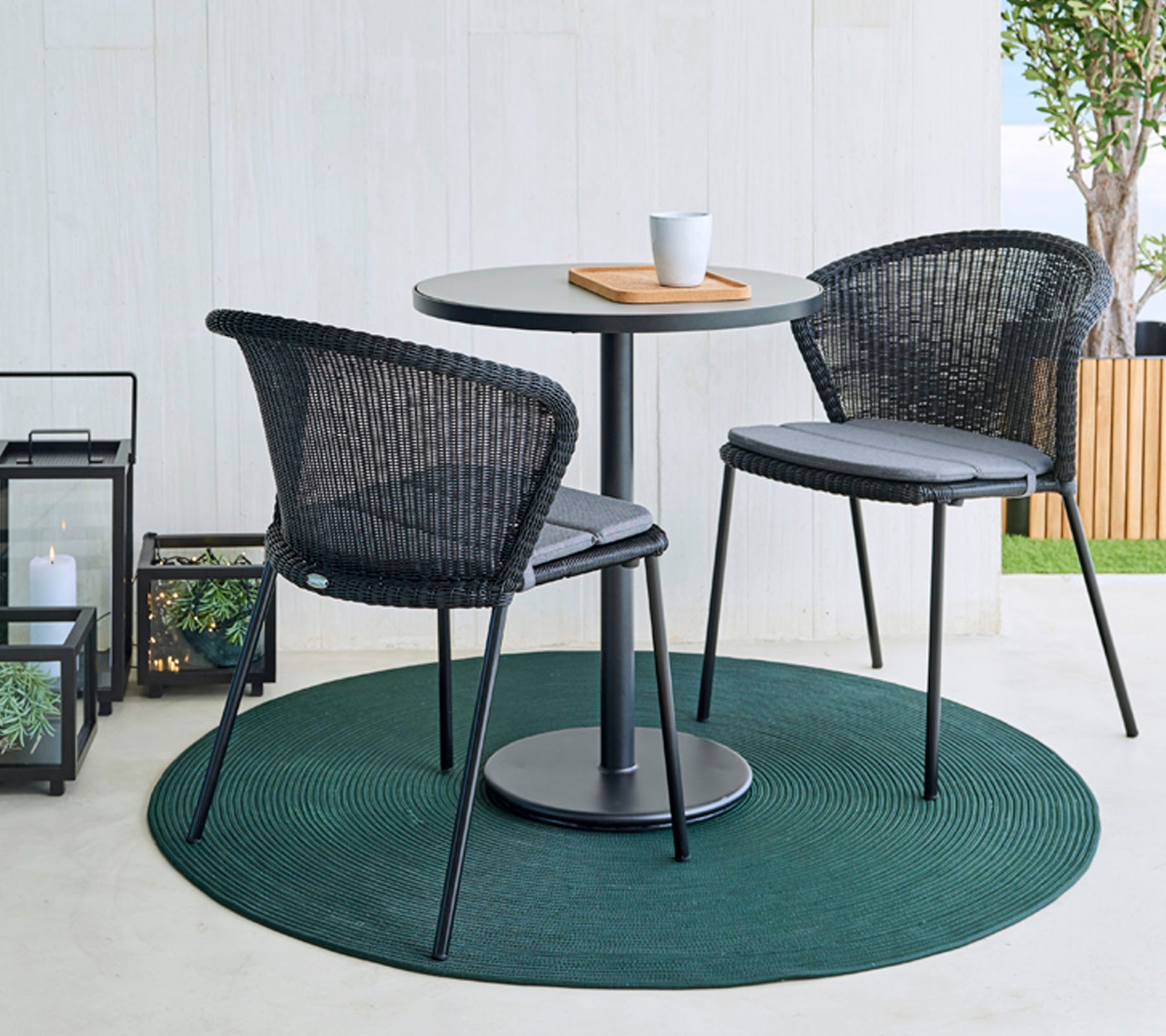 Une table ronde avec deux chaises en rotin noir sur un tapis vert, accompagnée d'une tasse à café et d'une planche en bois. Des lanternes décoratives en arrière-plan.