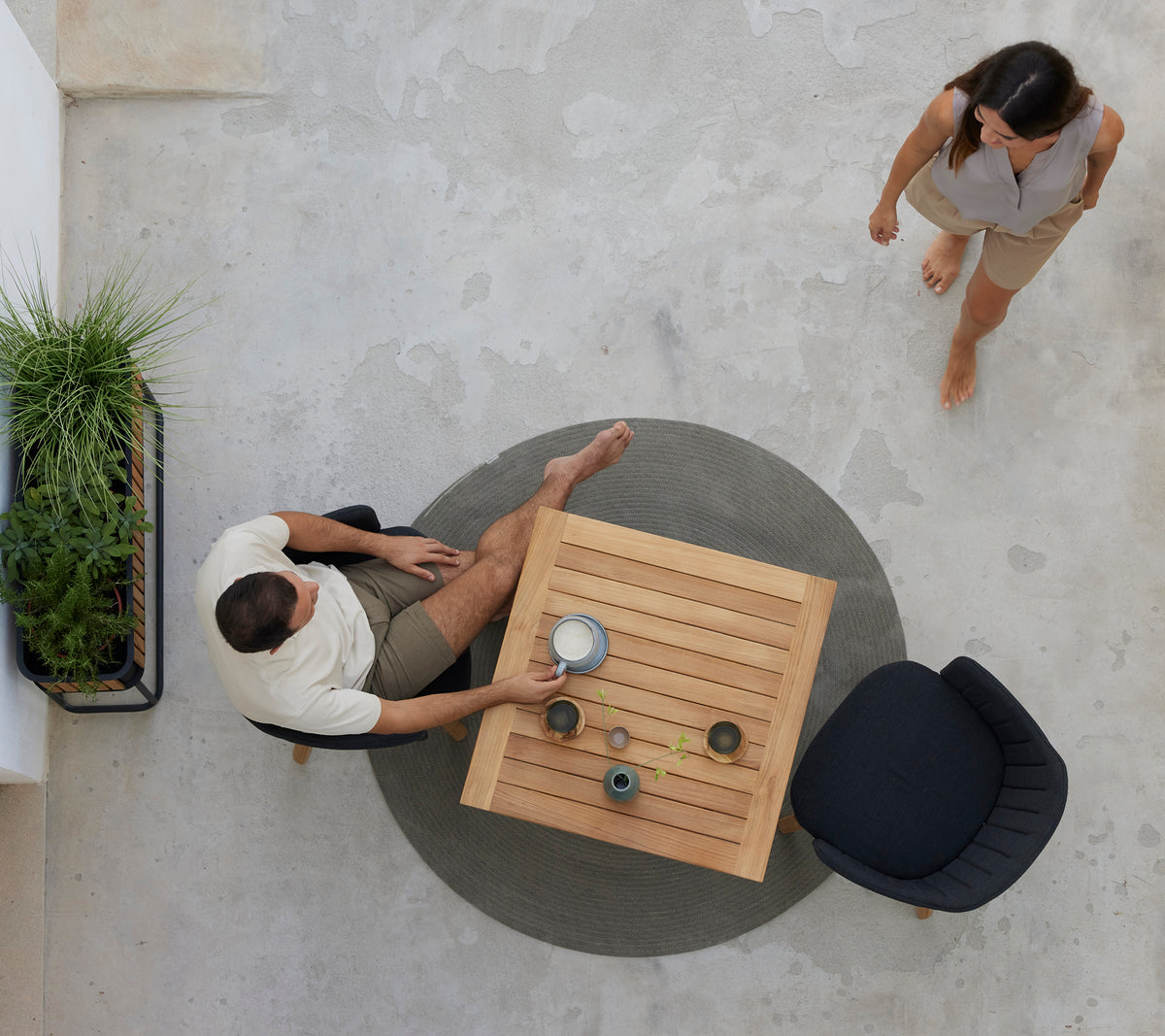 Deux personnes apprécient un café à une table en teck entourée de verdure, avec une chaise noire à proximité. Une atmosphère de café détendue est dépeinte.