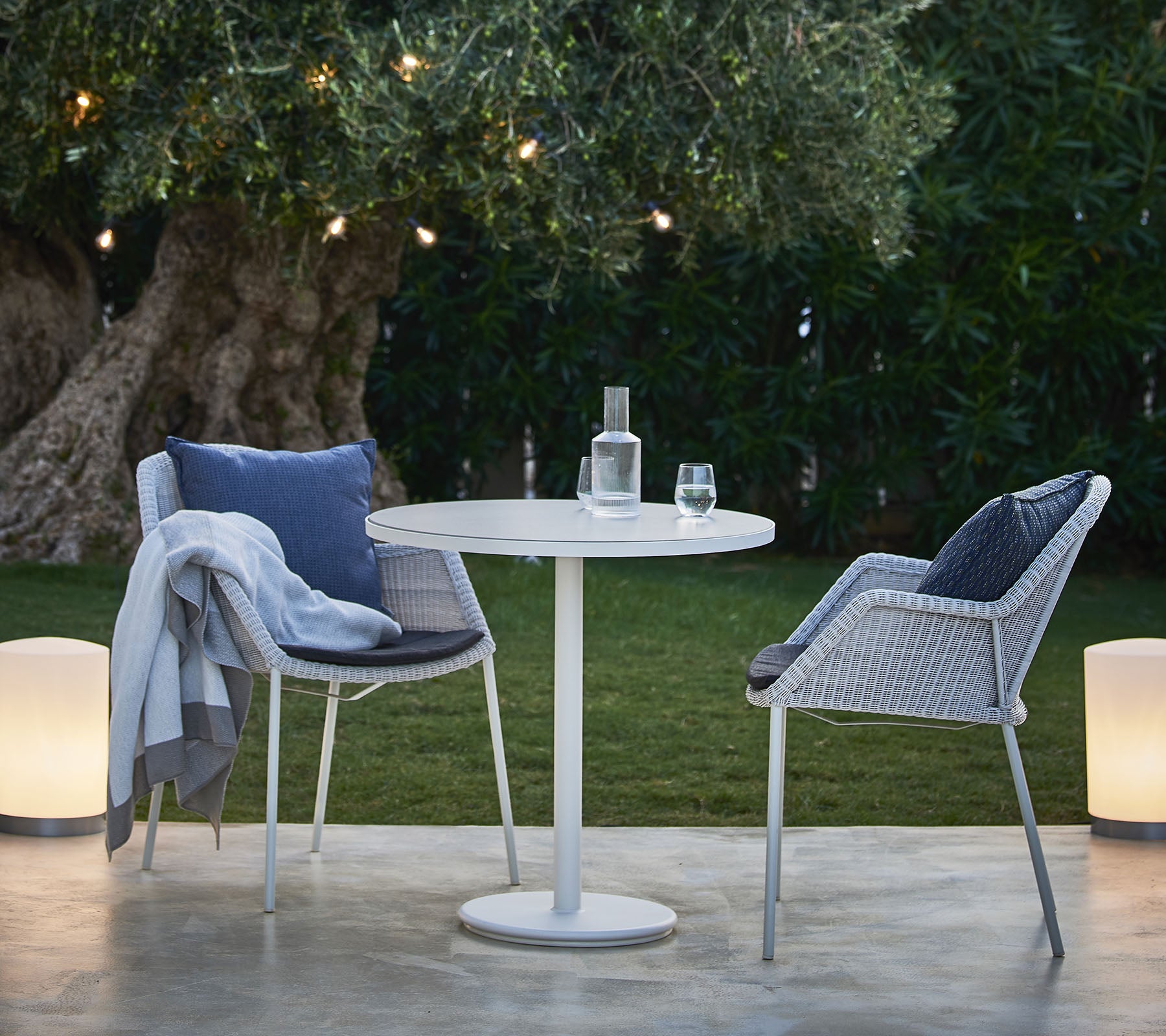 Une petite table ronde avec deux chaises élégantes, une bouteille d'eau et un verre, placée au milieu d'une verdure luxuriante et de lumières douces.