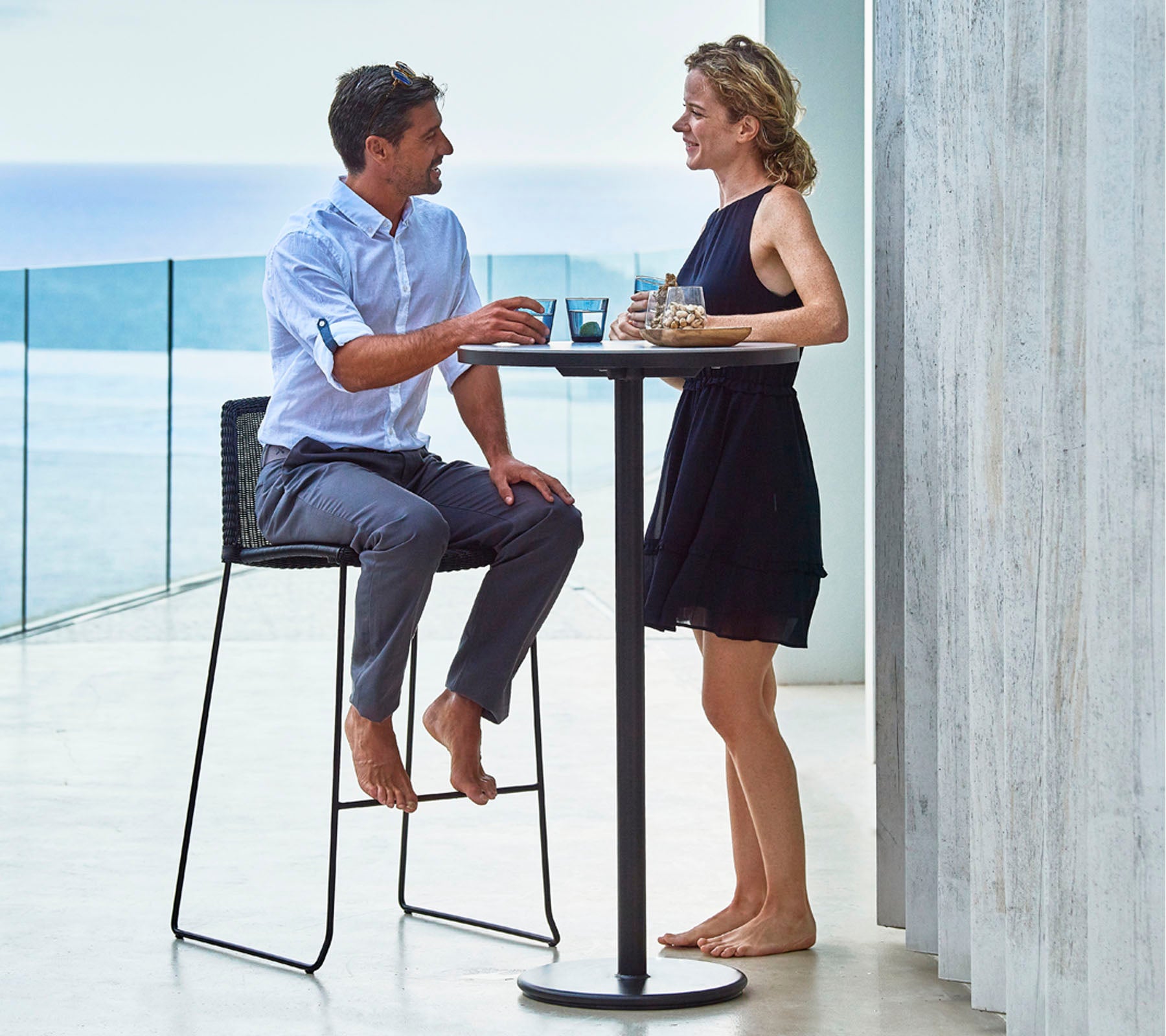 Un homme et une femme profitent de boissons à une table haute, engagés dans une conversation, avec un décor pittoresque d'eau au loin.