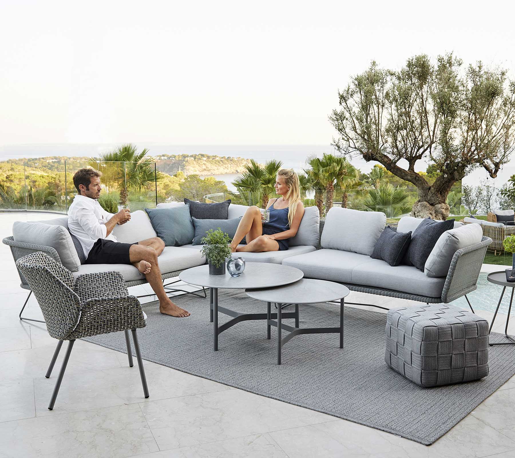 Une zone de sièges extérieure moderne avec un canapé, un fauteuil et une table basse, où deux personnes se détendent entourées de verdure et d'une vue pittoresque.
