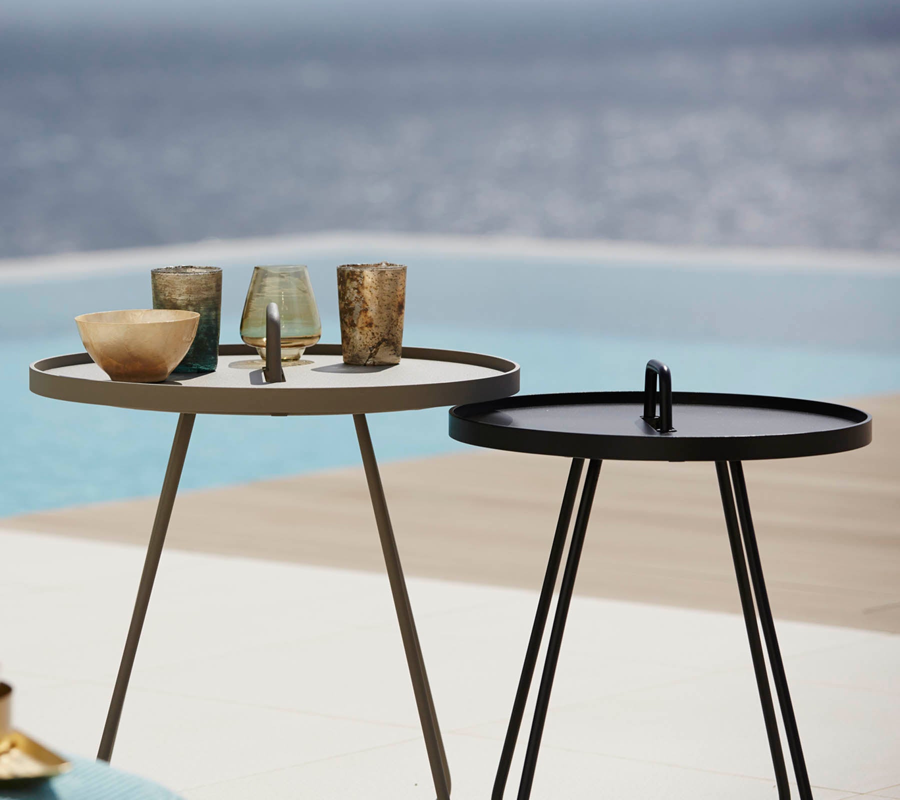Deux tables d'appoint élégantes avec divers objets en verre décoratif sur le dessus, placées près d'une paisible fontaine en arrière-plan.