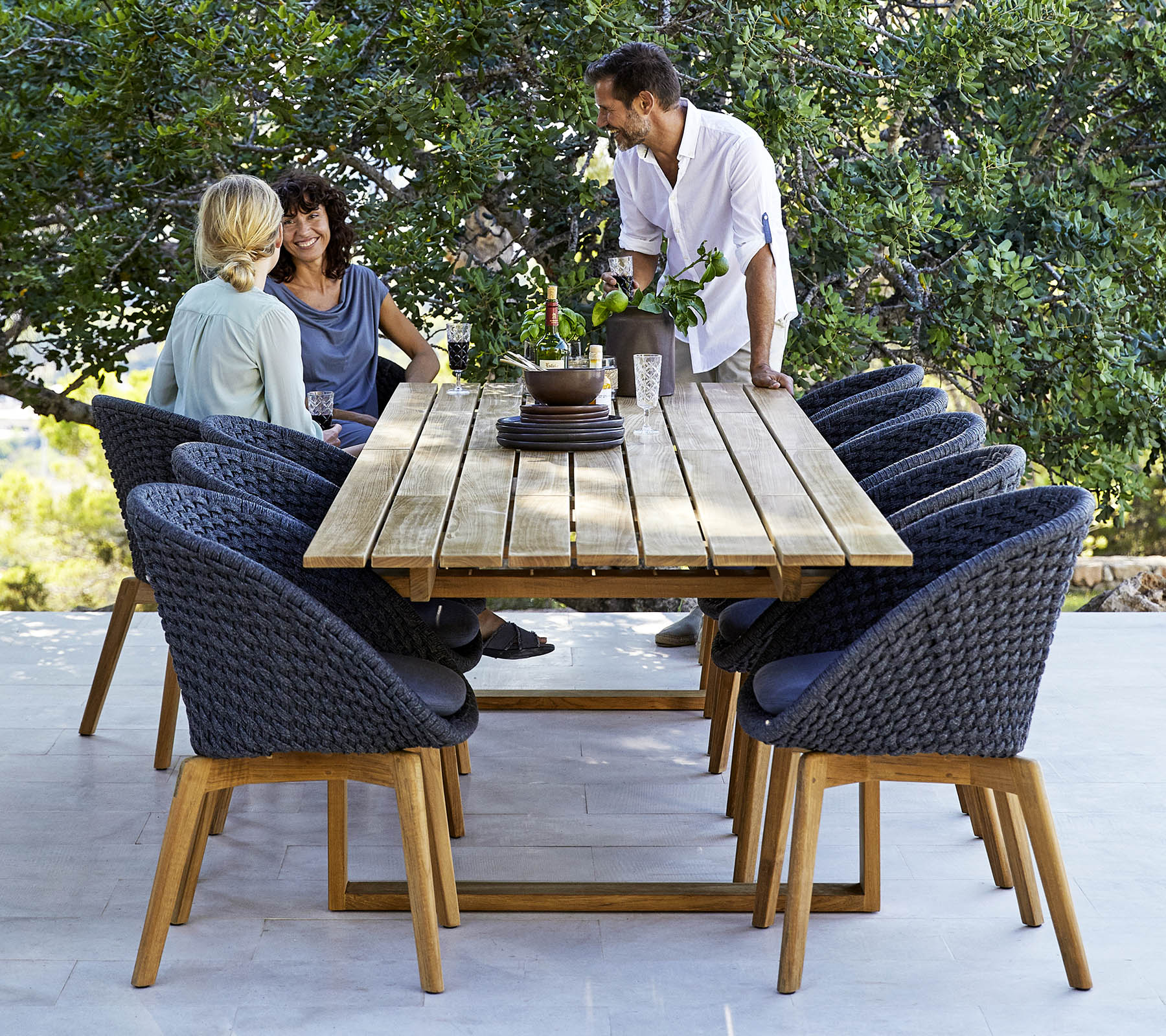 Une disposition de salle à manger élégante avec une table en bois entourée de chaises confortables, avec trois personnes engagées dans une conversation, au milieu de la verdure.