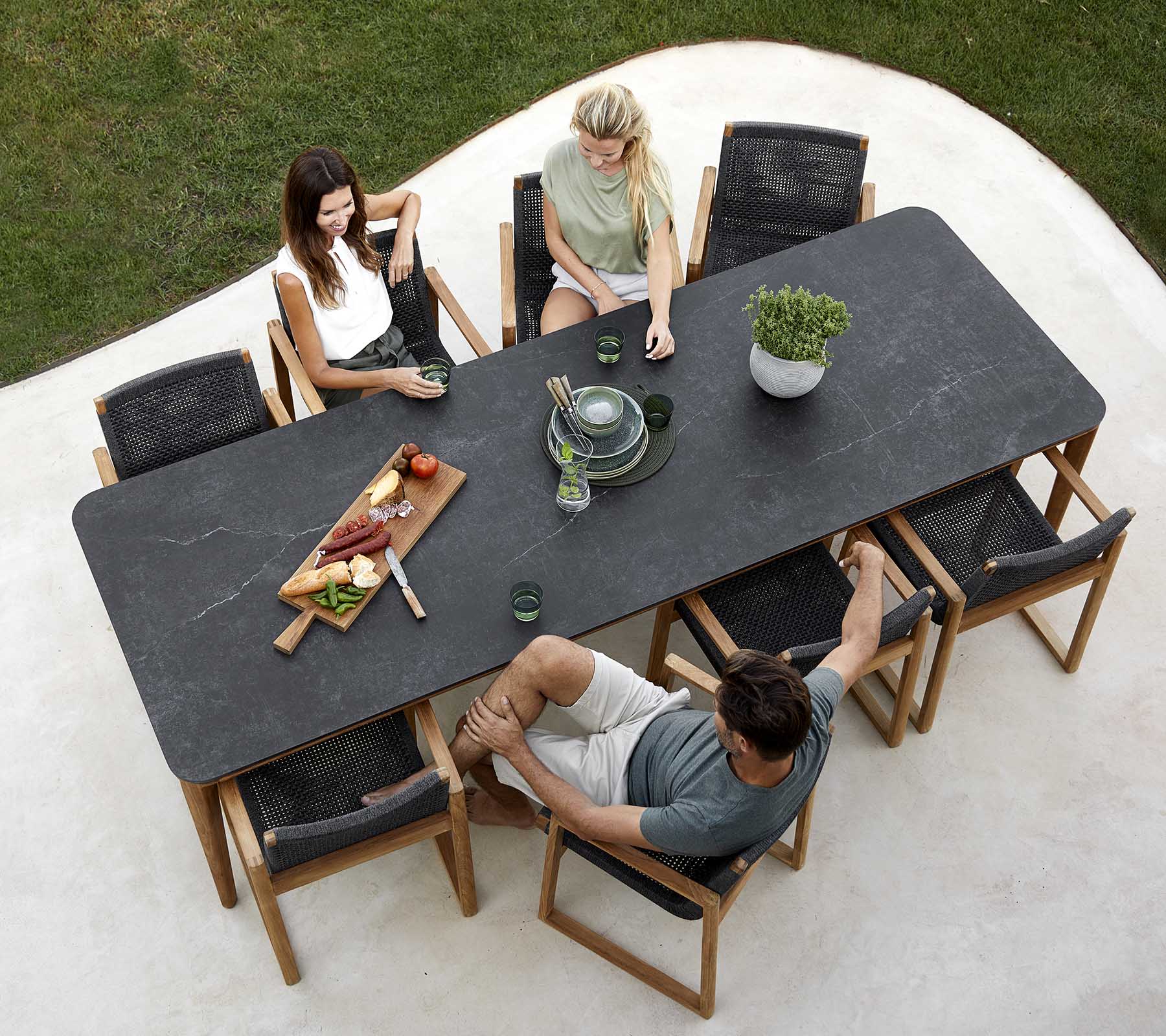 Un groupe de personnes apprécie un repas autour d'une grande table, avec des plats, des boissons et une plante en centre de table. Atmosphère décontractée.