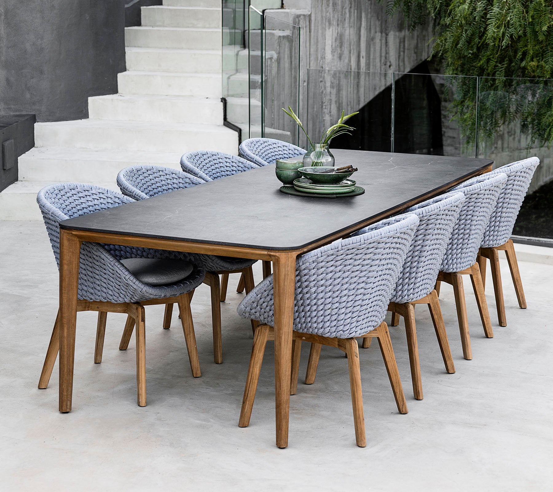 Une table à manger élégante avec une surface sombre, accompagnée de chaises tressées de couleur claire, complétée par des assiettes décoratives et du feuillage.