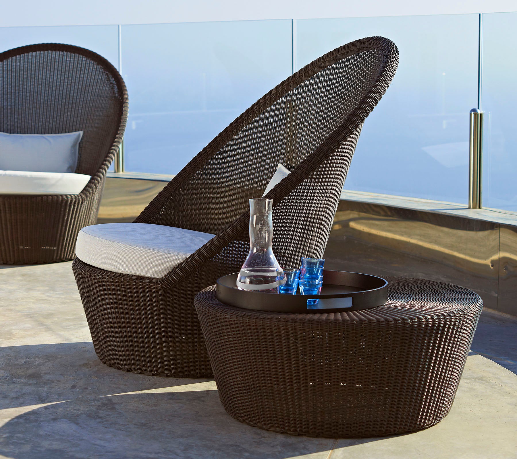 Chaise en rotin marron au design élégant avec un haut dossier et un coussin blanc, accompagnée d'une table d'appoint assortie et d'objets décoratifs.