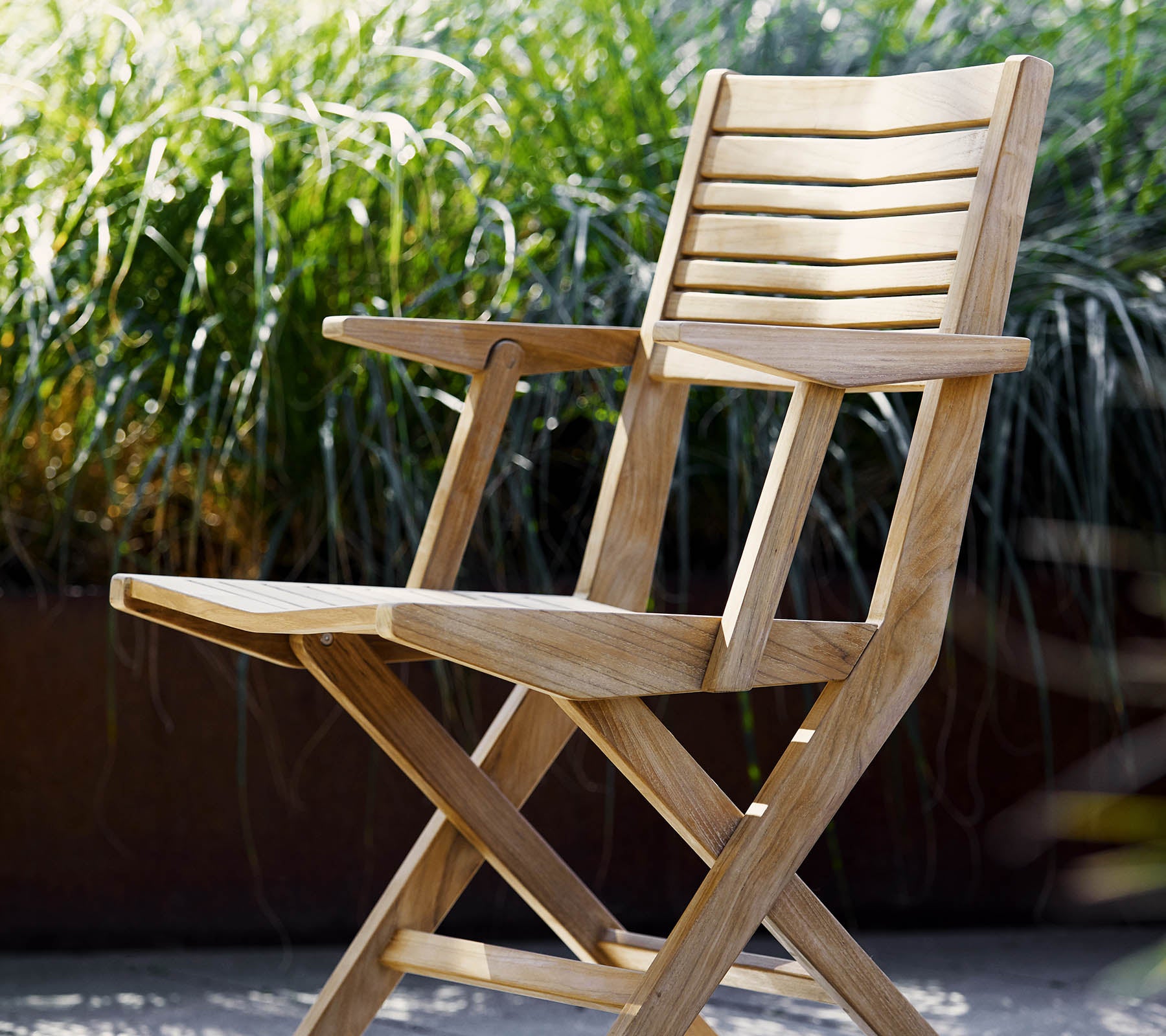 Une chaise pliante en bois avec des détails à lattes et des accoudoirs, entourée d'un feuillage vert luxuriant. Idéale pour se détendre et pour des espaces extérieurs.