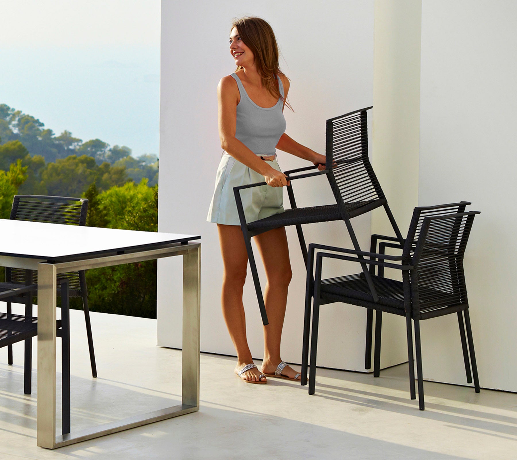 Une femme en débardeur léger et en shorts porte des chaises noires empilées à côté d'une table élégante, sur fond d'un arrière-plan naturel flou.