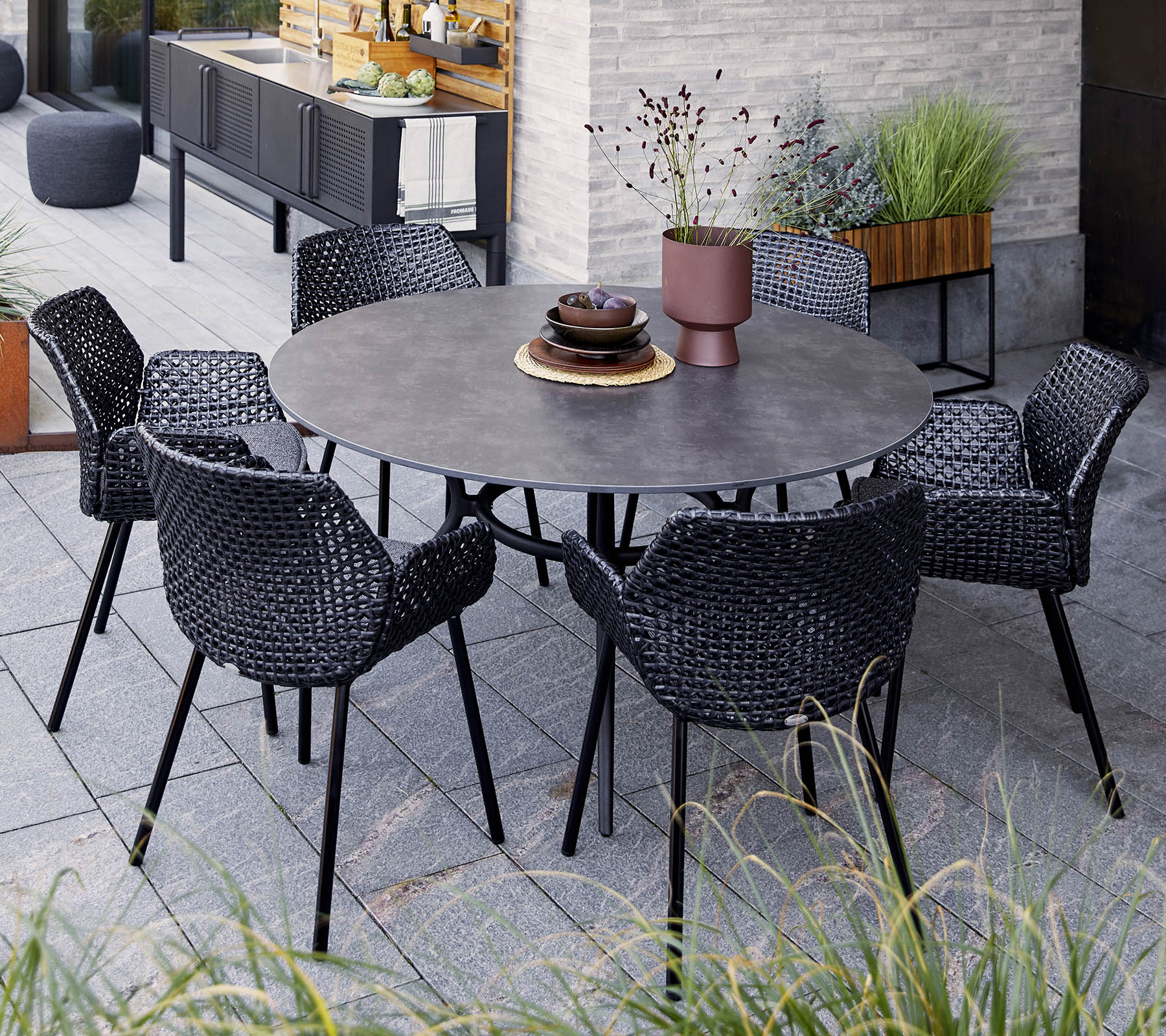 Table ronde avec six chaises noires tressées, ornée d'un bol décoratif et d'une plante en pot, mise en valeur parmi la verdure et un décor moderne.