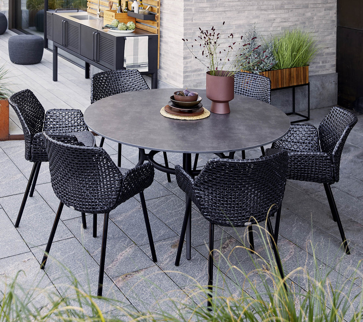 Table ronde avec six chaises noires tressées, ornée d'un bol décoratif et d'une plante en pot, mise en valeur parmi la verdure et un décor moderne.