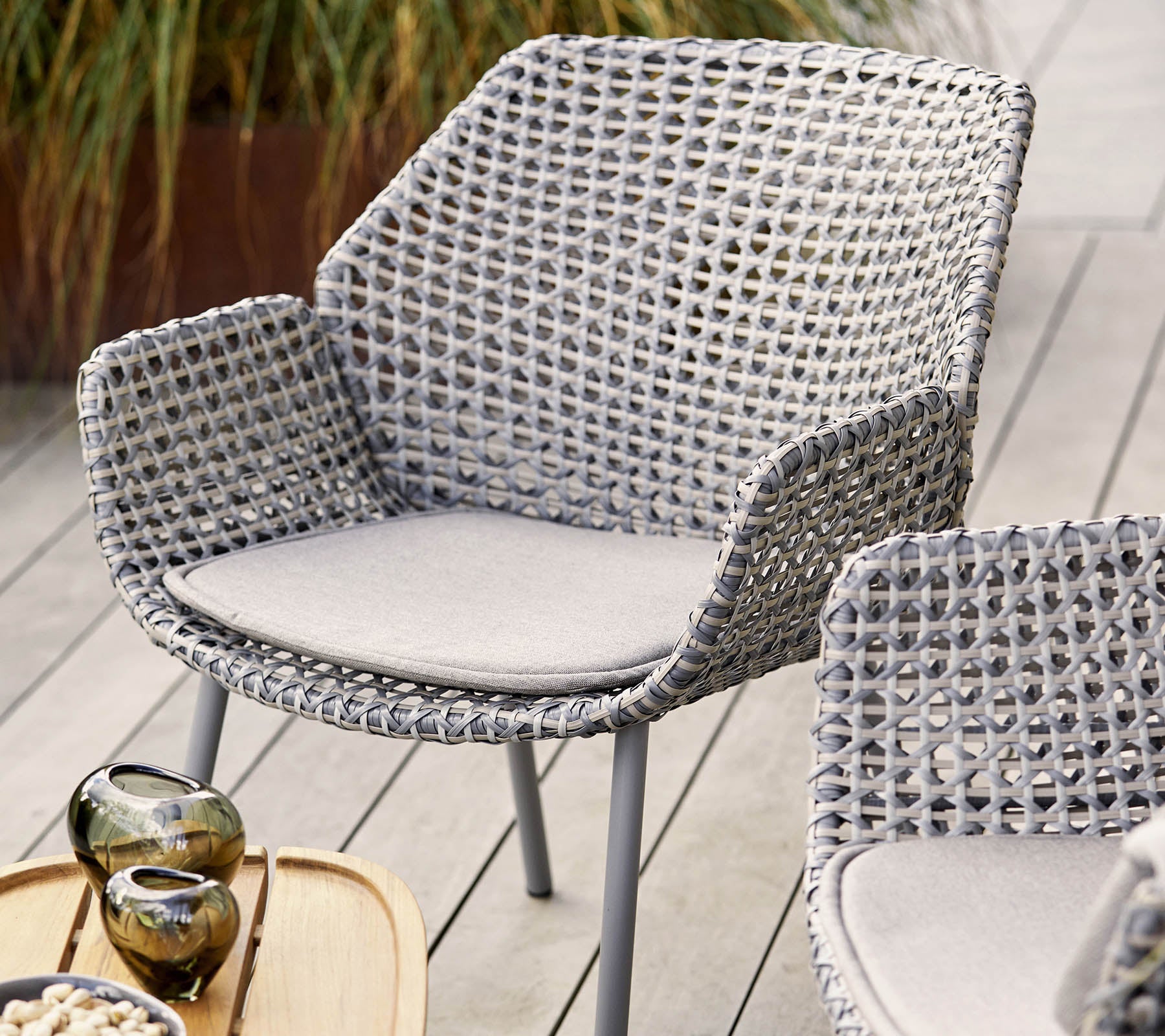 Chaise tissée élégante en gris clair avec un coussin confortable, complétée par des vases décoratifs et un plateau en bois.