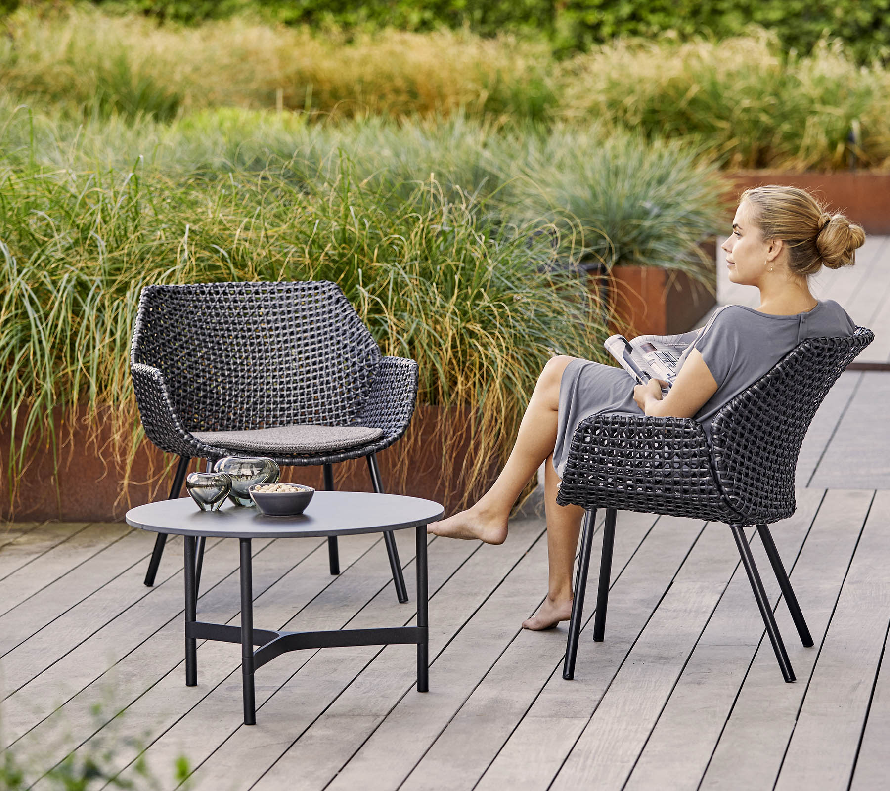 Deux chaises tressées élégantes avec une table assortie, entourées d'une végétation luxuriante. Une femme se détend en tenant un magazine.