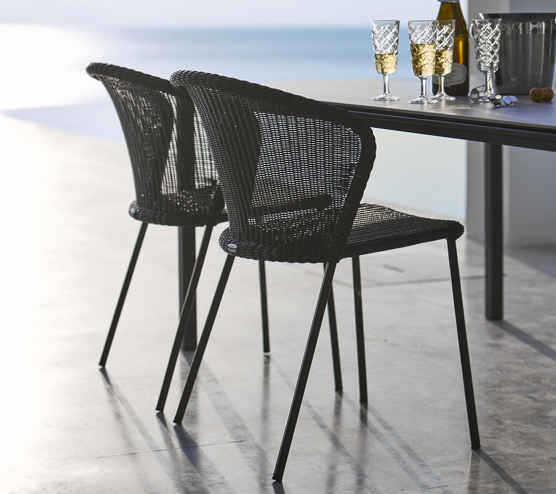 Deux chaises en rotin noires élégantes posées à côté d'une table garnie de verres et d'une bouteille, évoquant une atmosphère de repas décontractée.