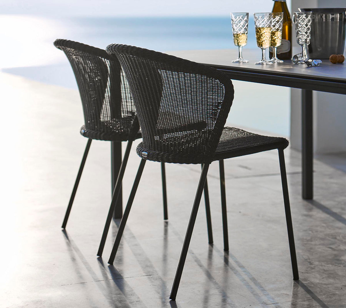Deux chaises en rotin noires élégantes posées à côté d'une table garnie de verres et d'une bouteille, évoquant une atmosphère de repas décontractée.