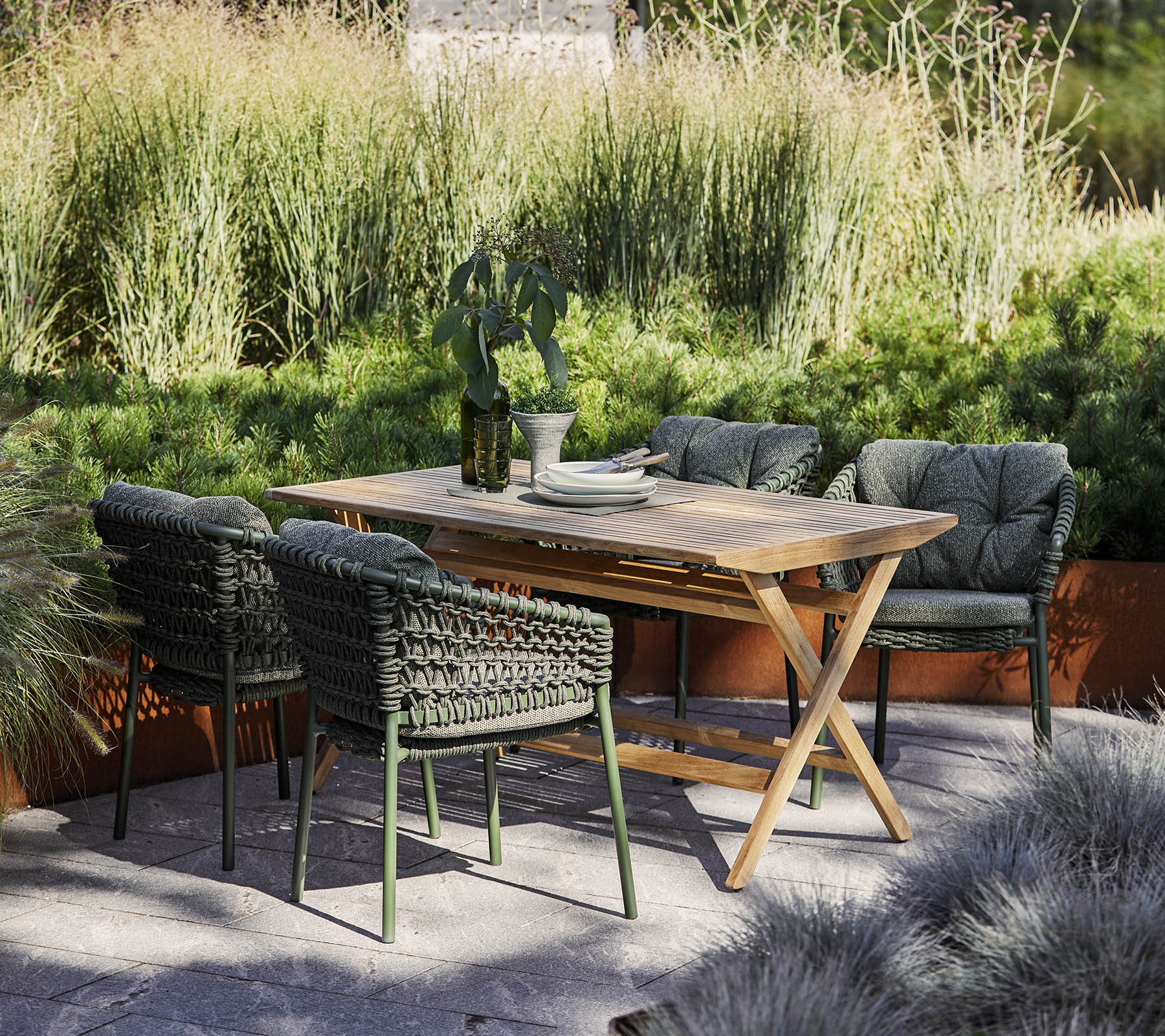 Un ensemble de salle à manger d'extérieur élégante avec une table en bois et quatre chaises en osier, entouré de verdure luxuriante et de plantes décoratives.