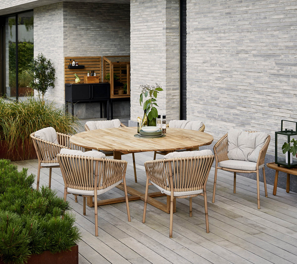 Table de salle à manger en bois rond entourée de chaises rembourrées, avec un centre de table décoratif en plante, contre un mur en pierre texturé.