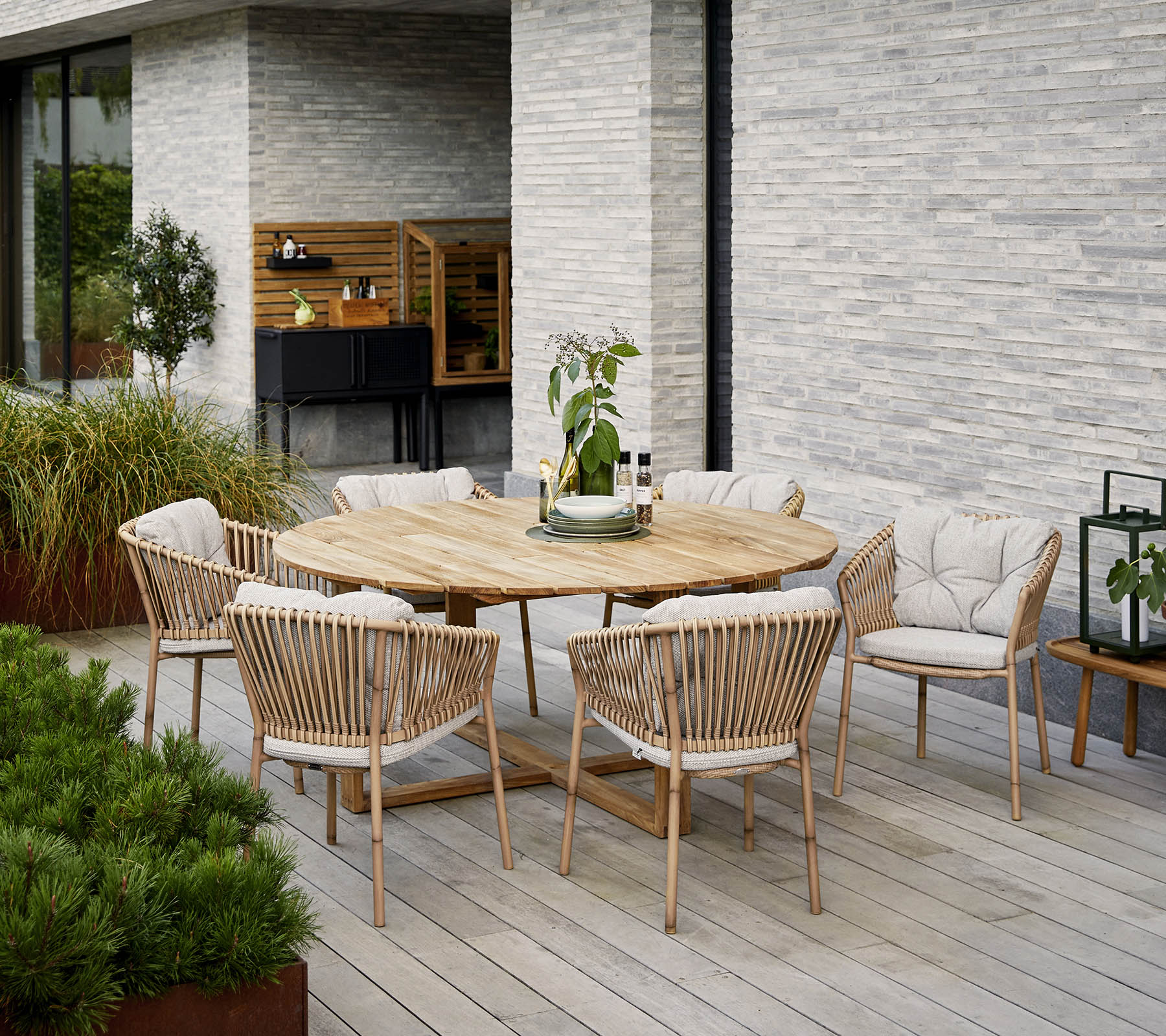 Table de salle à manger en bois rond entourée de chaises rembourrées, avec un centre de table décoratif en plante, contre un mur en pierre texturé.