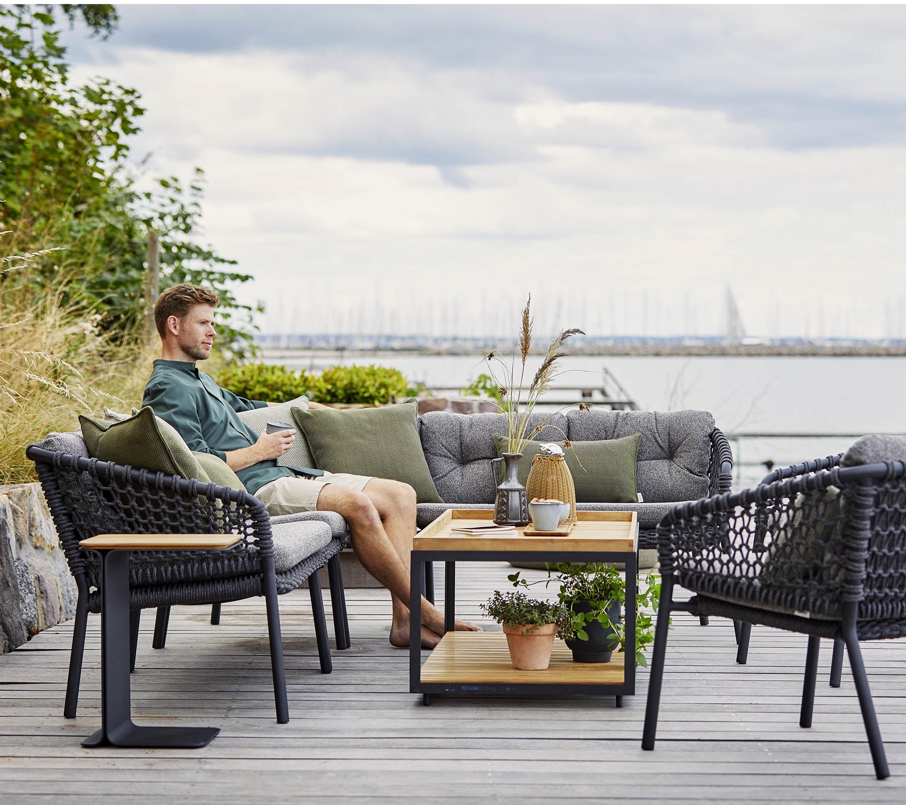Un coin salon confortable avec un homme se relaxant, comportant des chaises en rotin élégantes et une table basse en bois ornée de plantes et d'objets décoratifs.