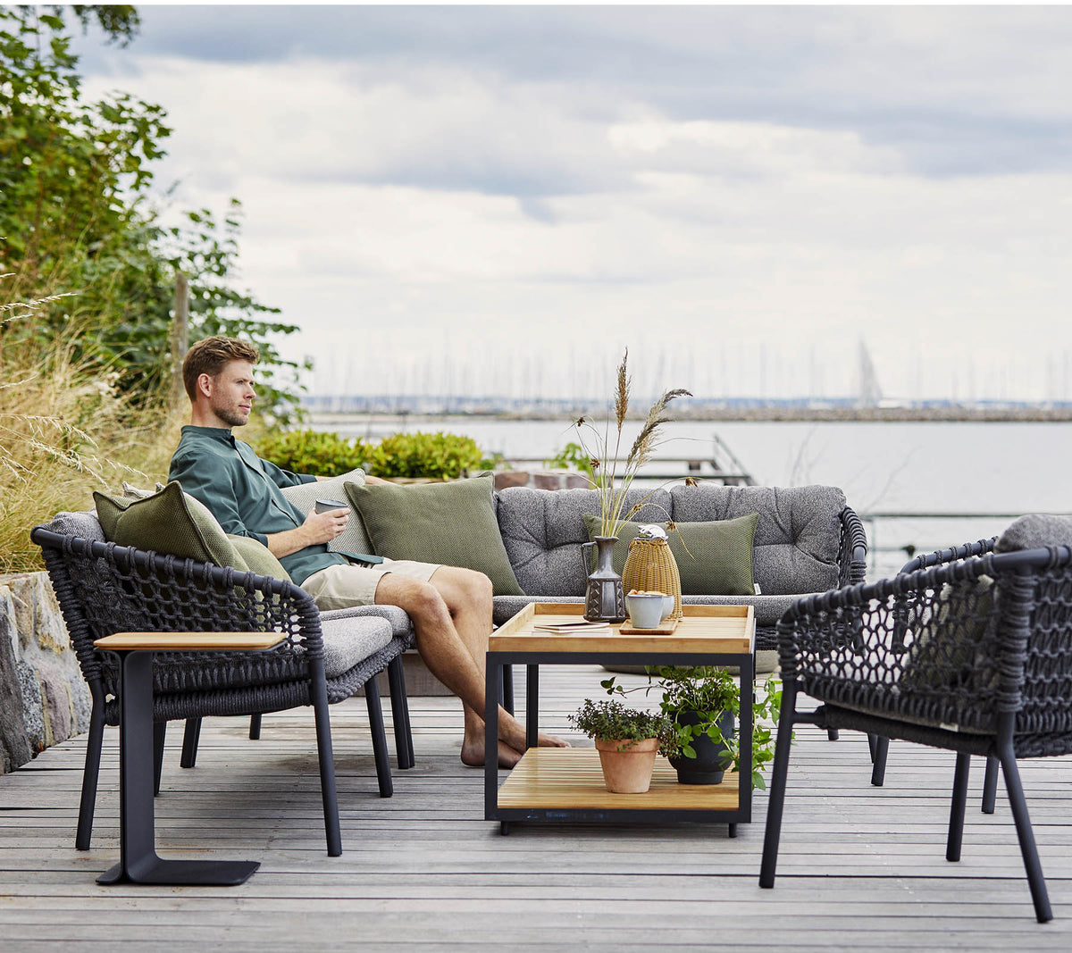 Un coin salon confortable avec un homme se relaxant, comportant des chaises en rotin élégantes et une table basse en bois ornée de plantes et d'objets décoratifs.