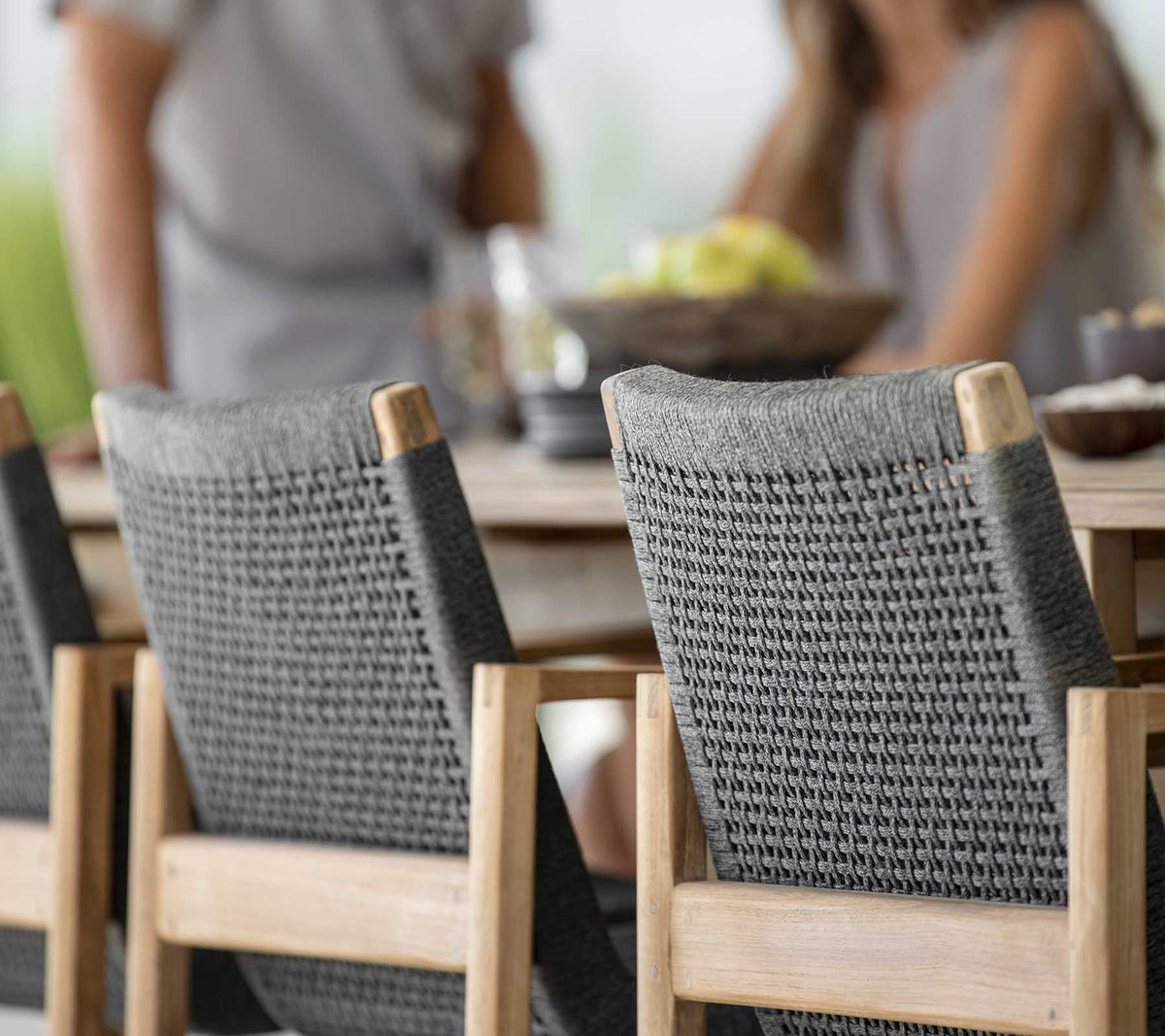 Scène de repas en plein air avec une table en bois et deux chaises tissées grises élégantes, où des gens partagent un repas ensemble.