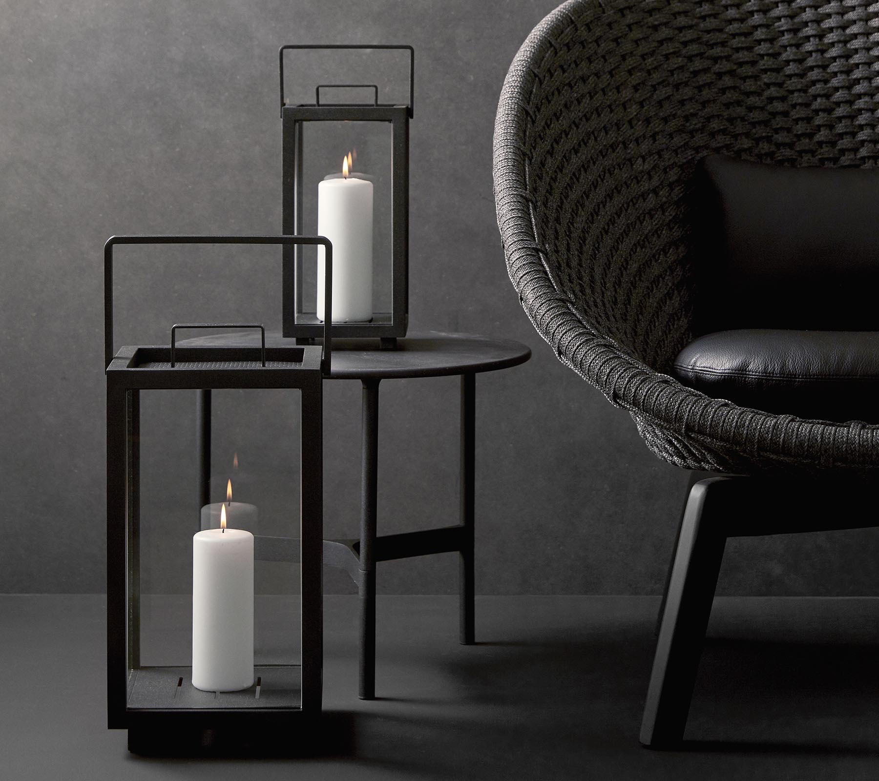 Deux lanternes élégantes avec des bougies blanches, accompagnées d'une chaise moderne et texturée sur une table minimaliste contre un fond sombre.