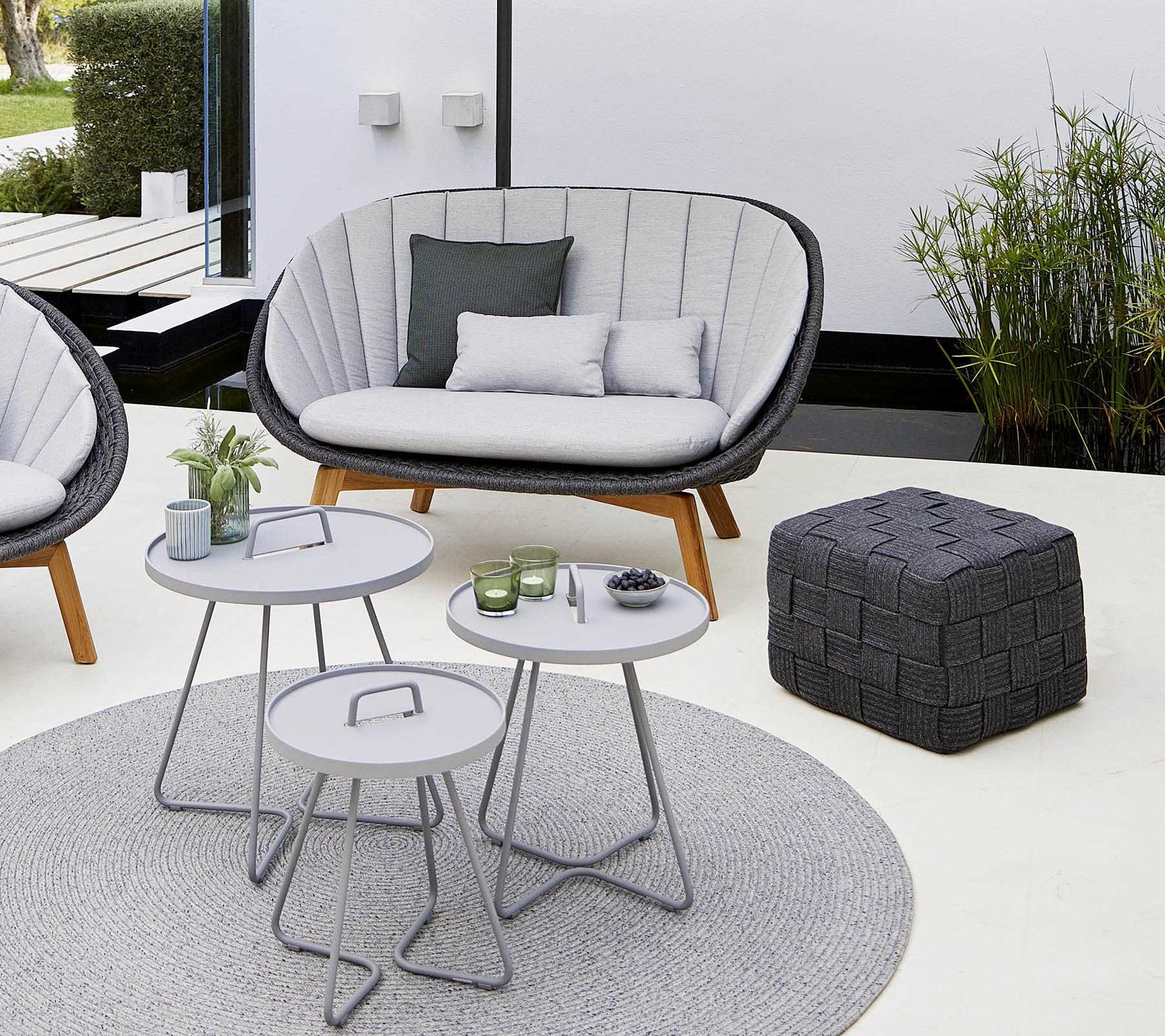 Espace salon confortable avec un canapé élégant, quelques tables rondes, un pouf décoratif et un tapis doux, entouré de verdure.