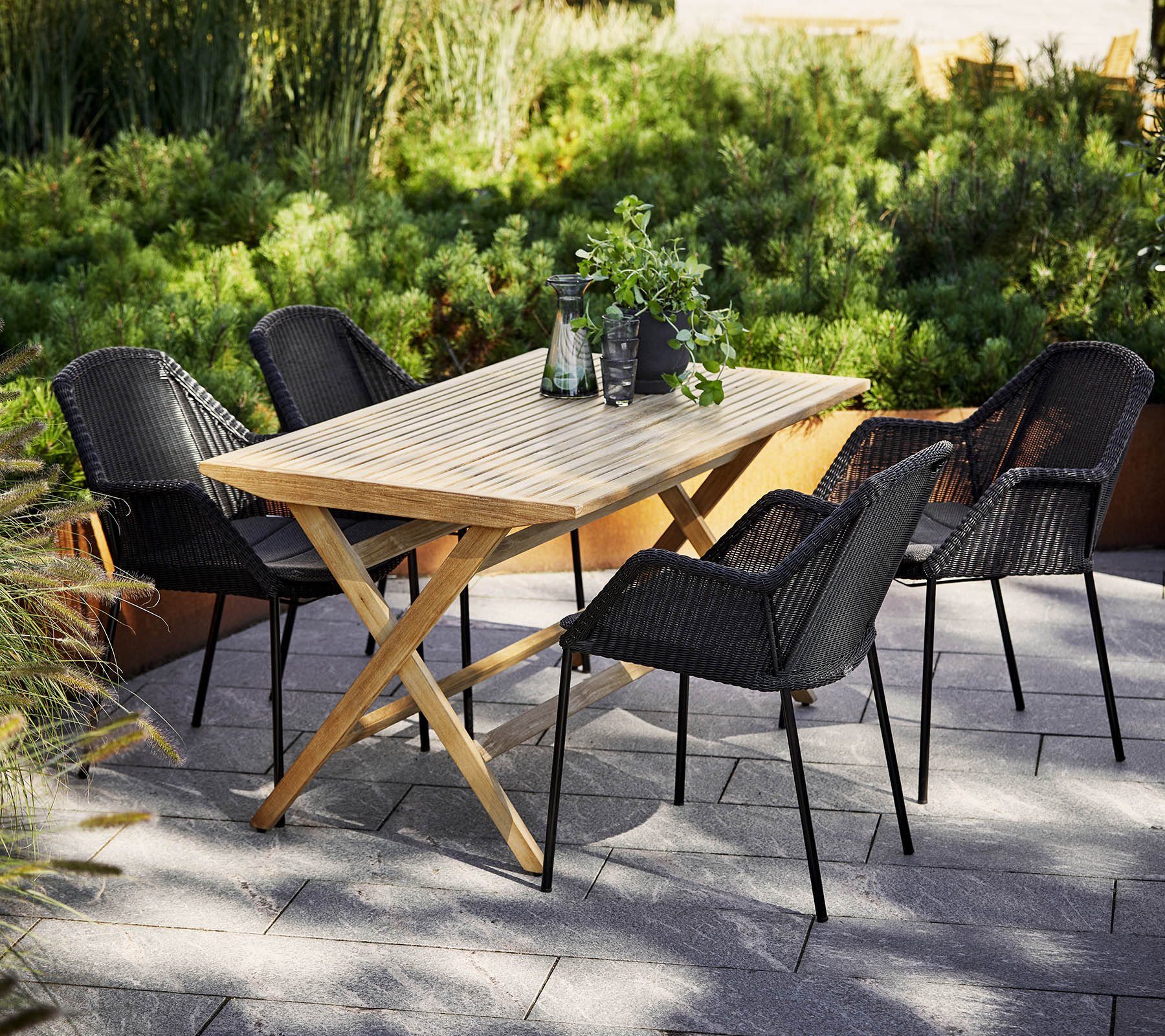 Une table à manger en bois entourée de quatre chaises tressées noires, avec une végétation en arrière-plan et des objets décoratifs sur la table.
