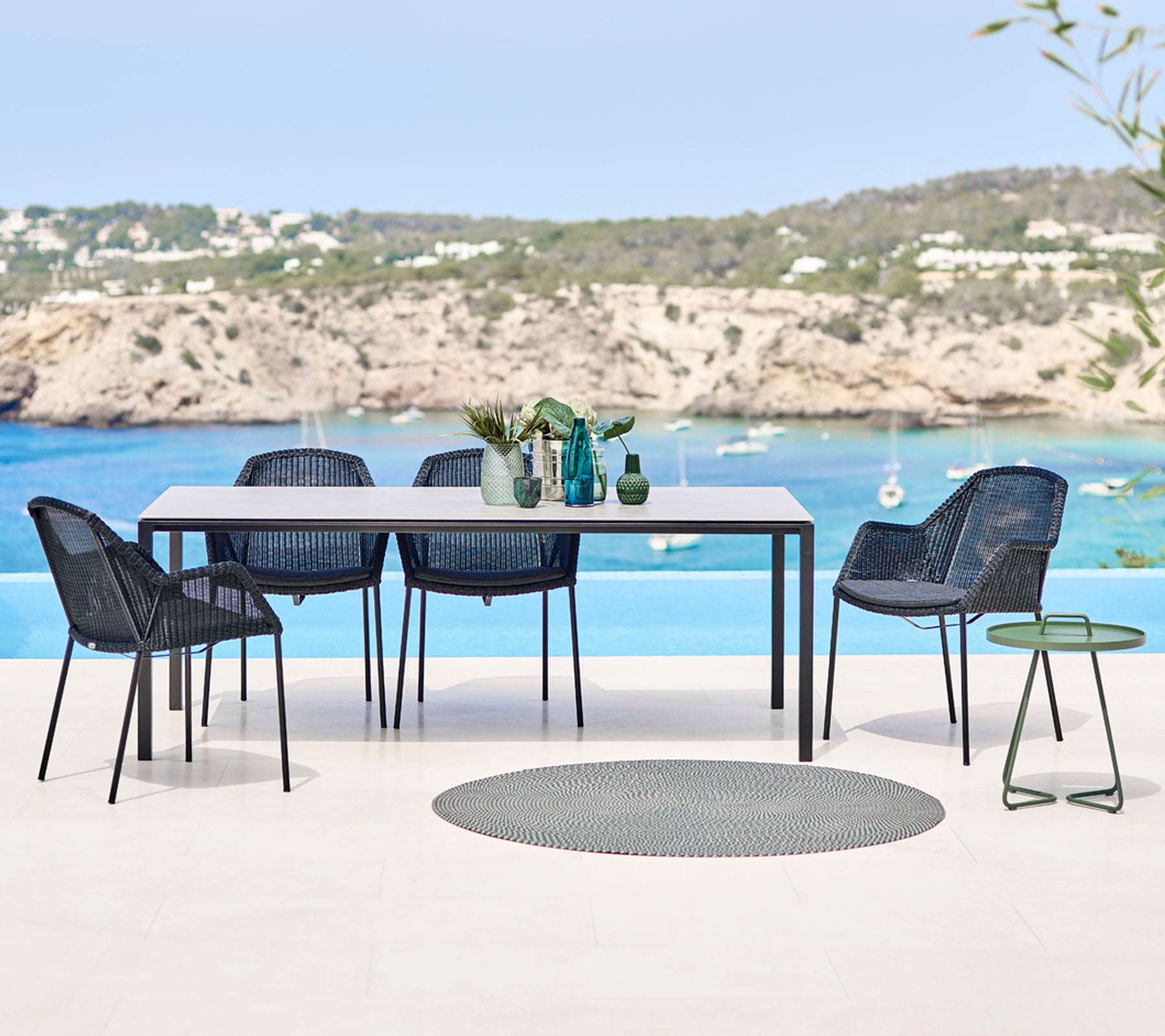 Un ensemble de salle à manger d'extérieur moderne comprenant une table rectangulaire, des chaises en osier et une petite table d'appoint, avec une toile de fond de plage pittoresque.