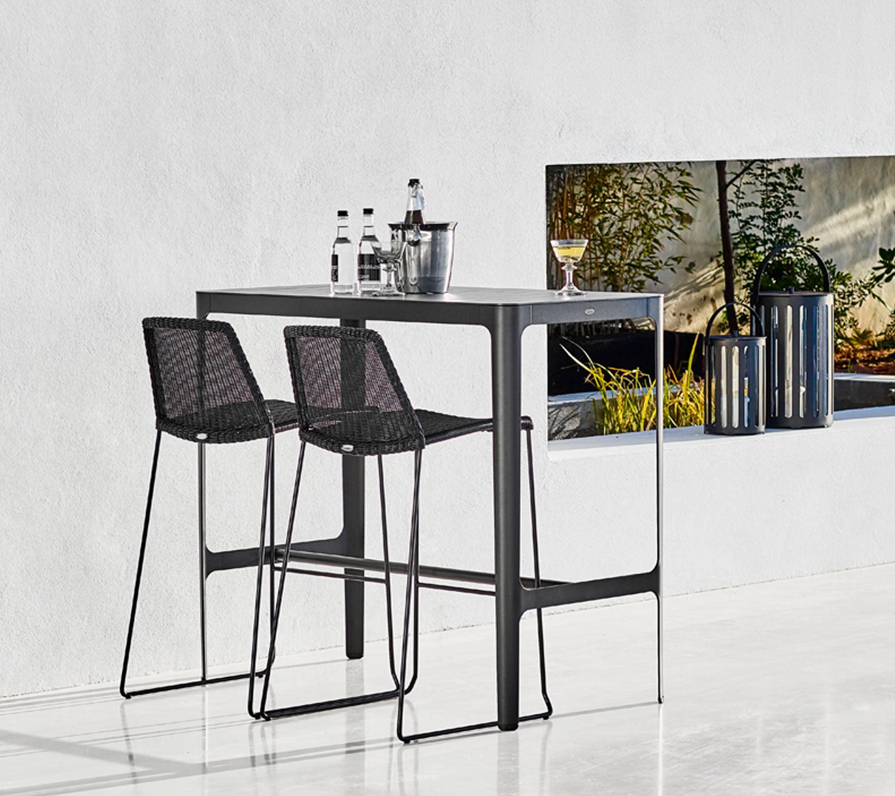 Table de bar élégante avec deux tabourets tressés noirs, accompagnée de verres et d'un seau stylé, sur fond minimaliste.