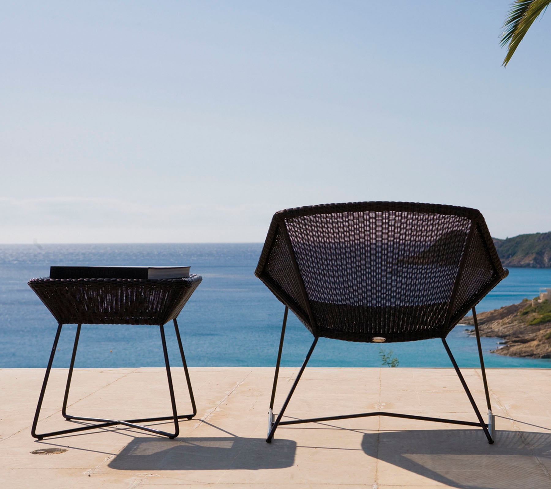 Deux chaises d'extérieur modernes et noires avec un design géométrique sont positionnées à côté d'une table, surplombant une mer calme sous un ciel dégagé.