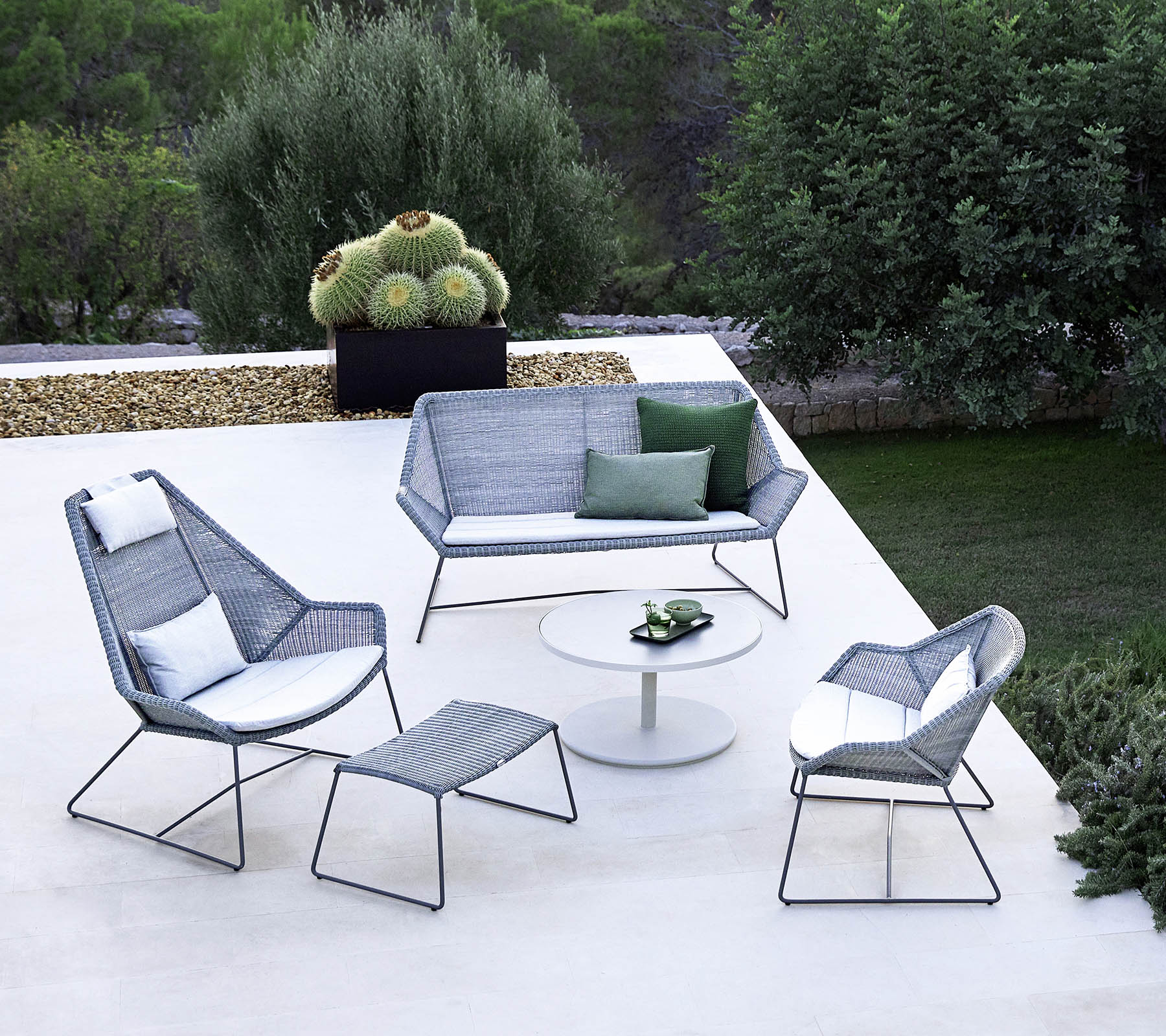 Aménagement de sièges extérieur élégant avec un canapé, deux fauteuils, un repose-pieds et une table basse, complété par de la verdure en arrière-plan.