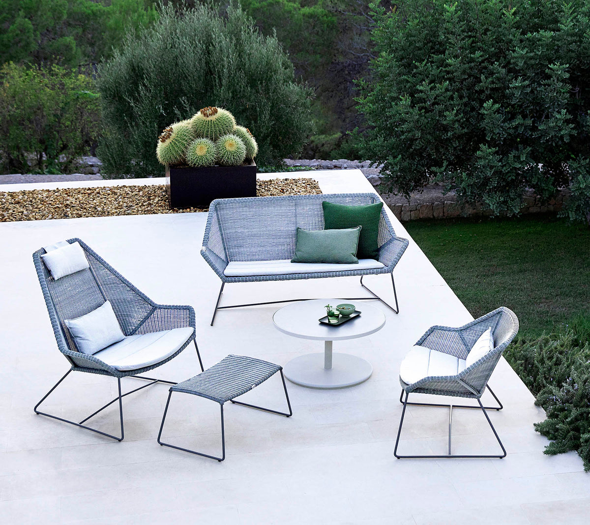 Aménagement de sièges extérieur élégant avec un canapé, deux fauteuils, un repose-pieds et une table basse, complété par de la verdure en arrière-plan.