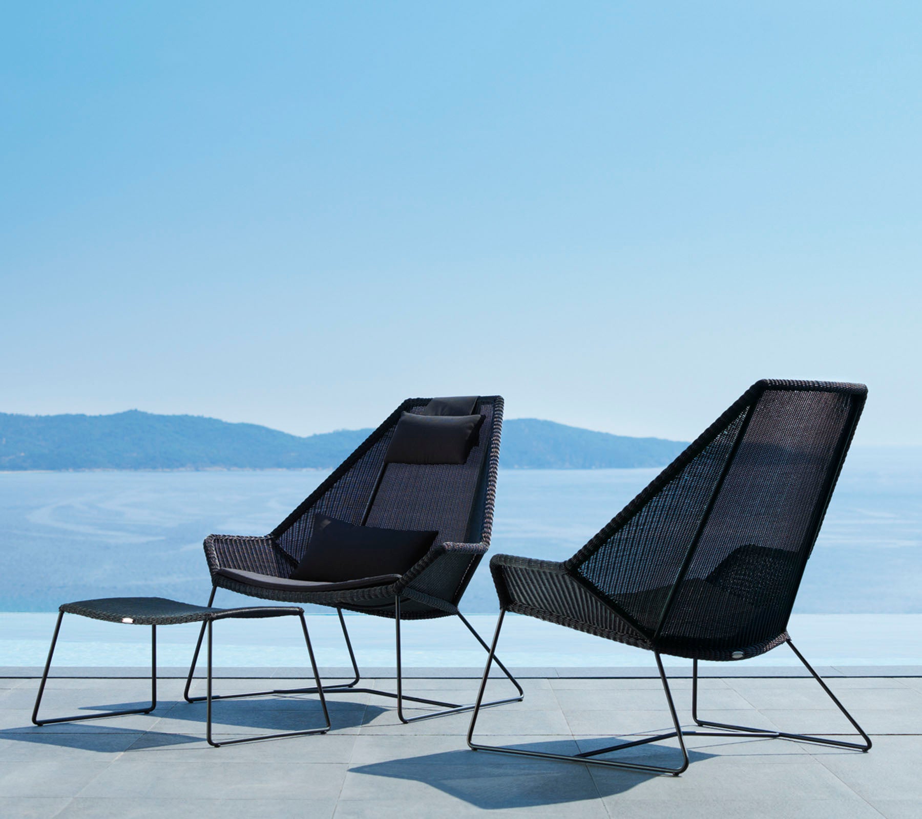 Deux chaises modernes et élégantes aux lignes épurées et au design en maille ouverte, accompagnées d'un repose-pieds assorti, offrant une vue sur une eau sereine.