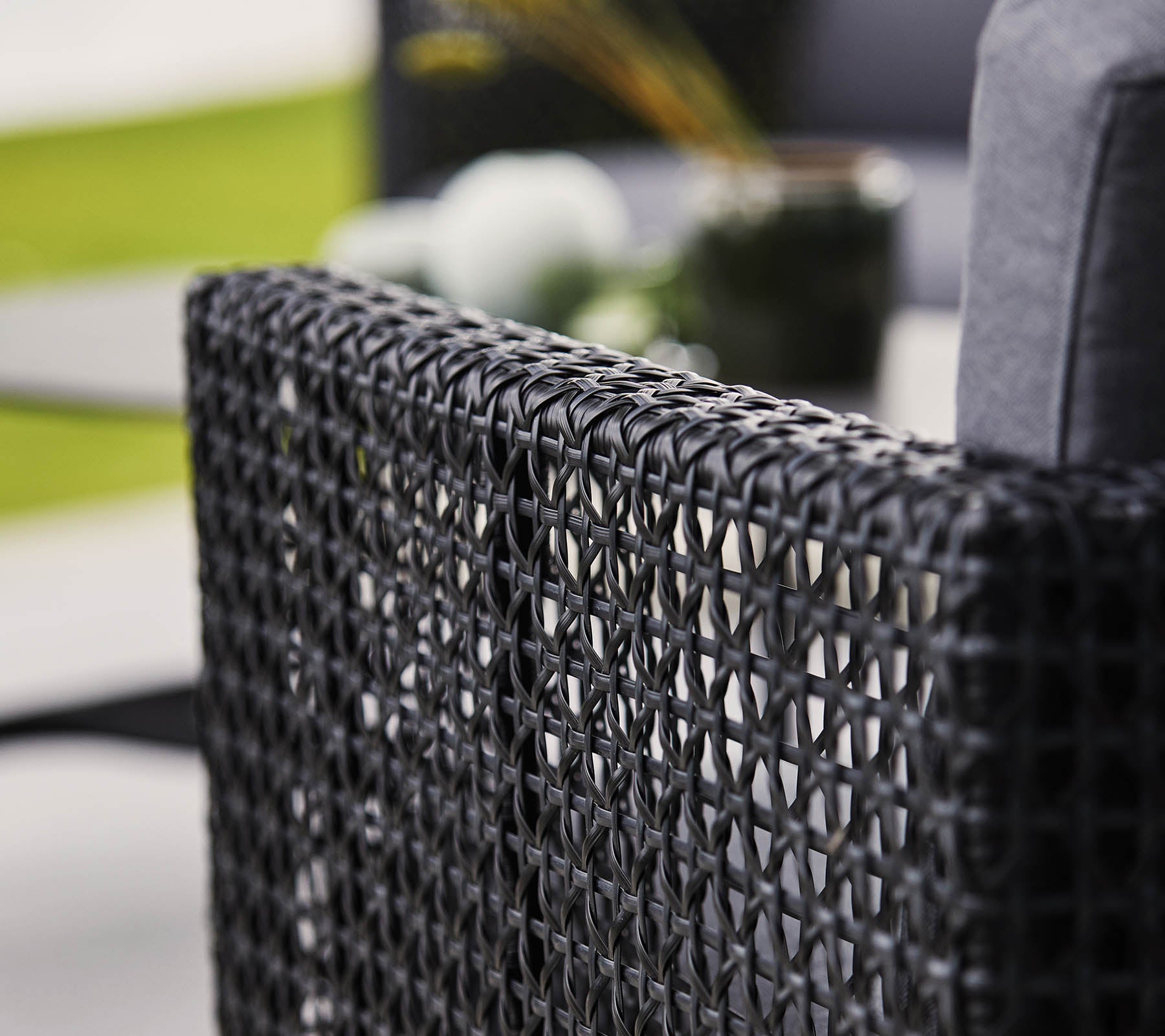 Dossier de chaise noir tressé avec un coussin doux, présentant des éléments décoratifs flous en arrière-plan.