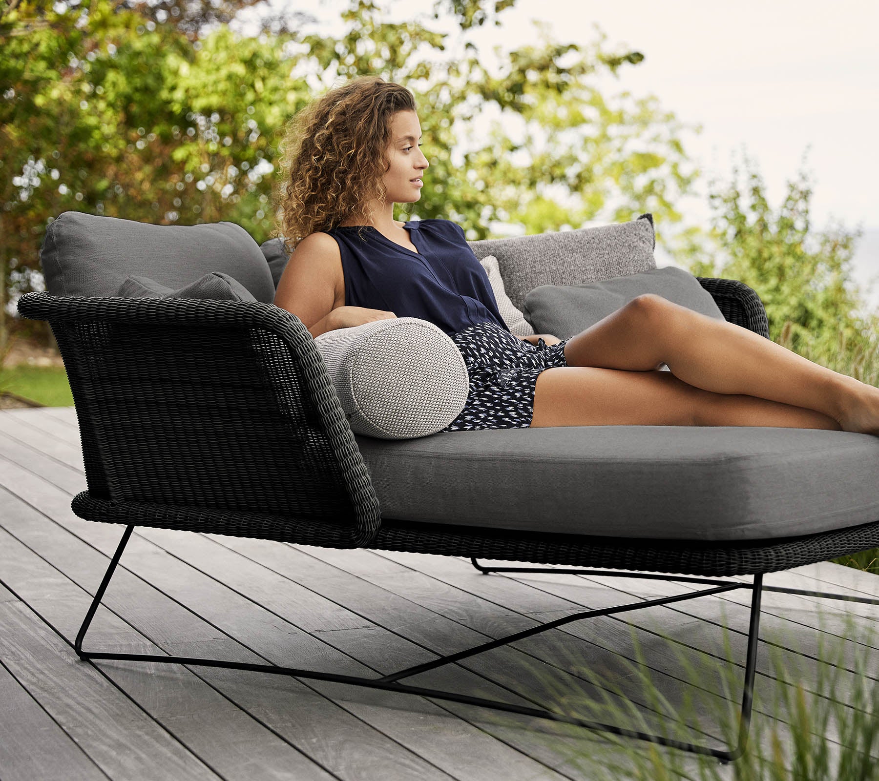 Une femme se détend sur une chaise longue élégante et rembourrée ornée de coussins, entourée de verdure.