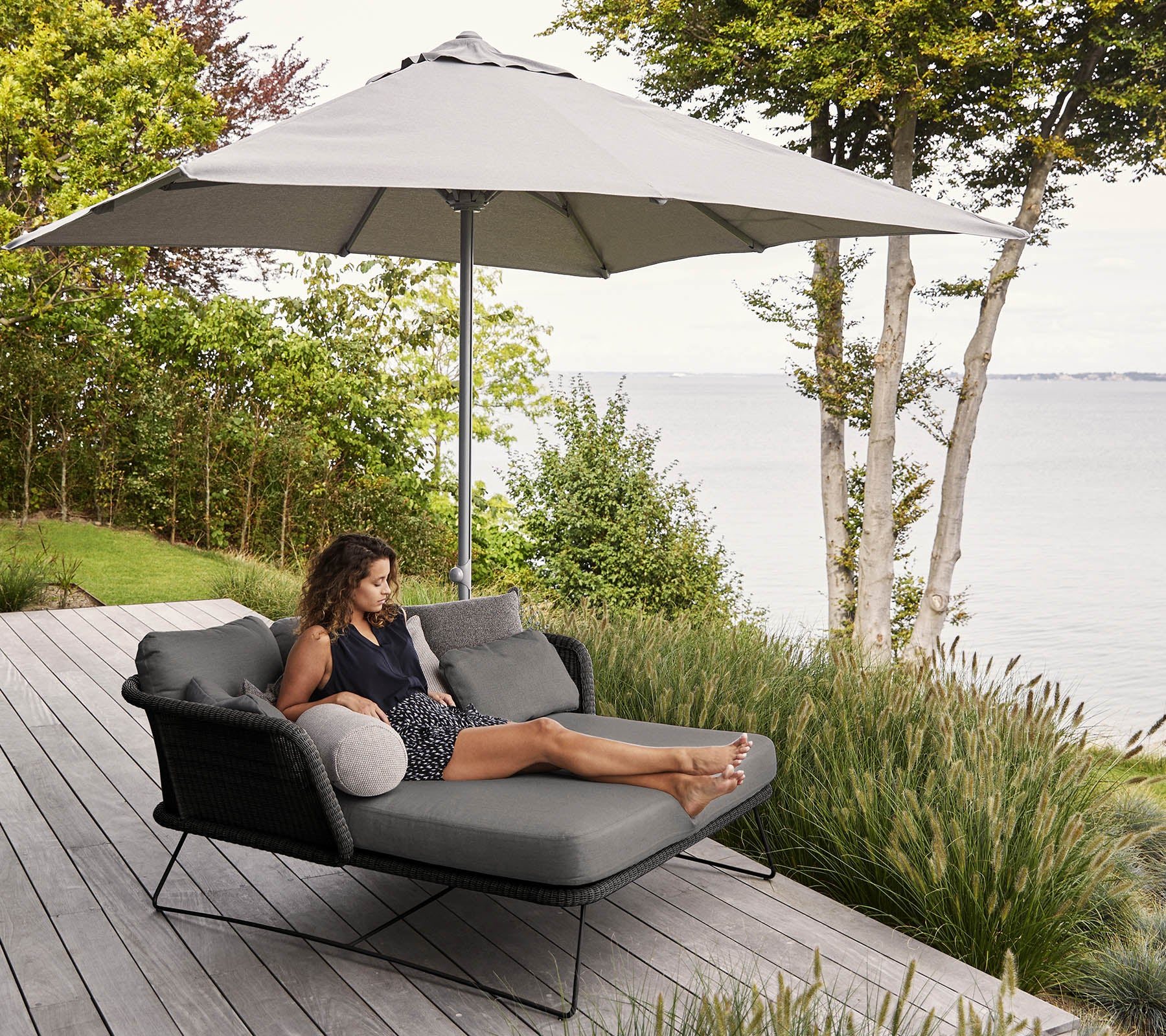Une femme se détend sur un fauteuil moderne avec un grand parasol, entourée de verdure et offrant une vue pittoresque sur le front de mer.