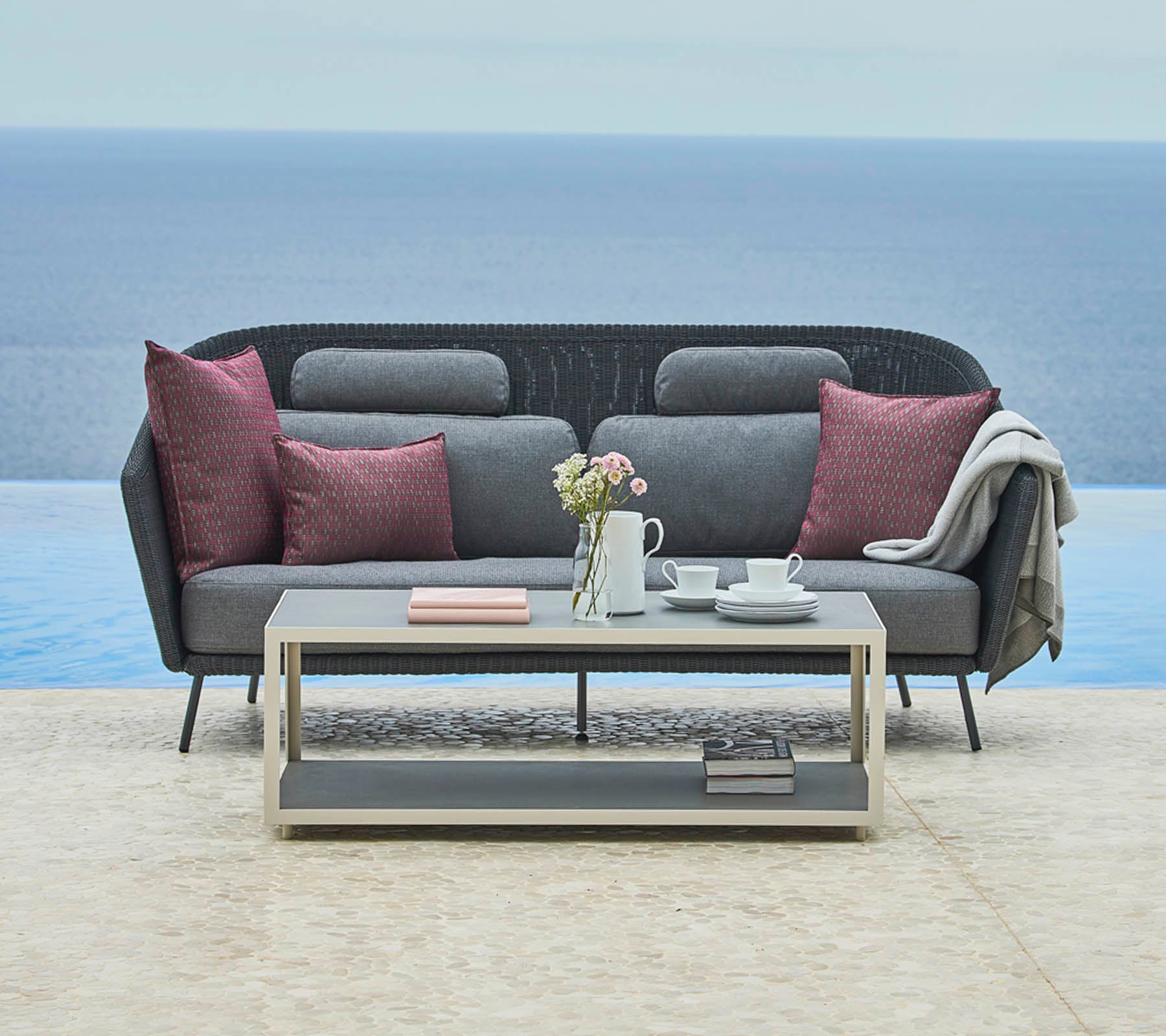 Canapé gris élégant avec des coussins décoratifs et un plaid confortable, complété par une table basse épurée avec une théière et des livres, devant un paysage serein.