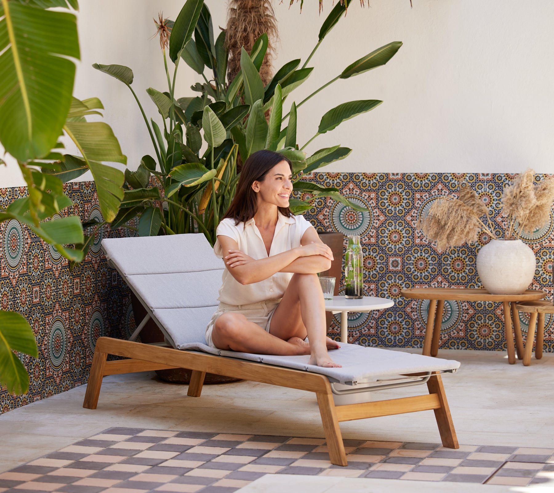 Une femme se détend sur une chaise longue entourée de plantes vibrantes et de décorations à motifs, appréciant une boisson rafraîchissante.