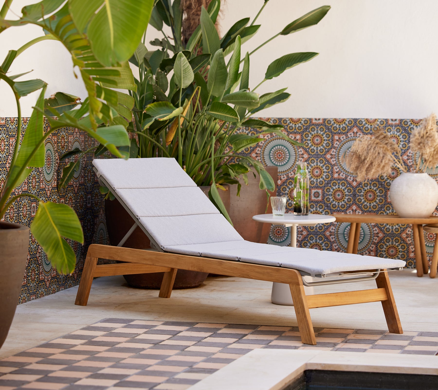 Un fauteuil moderne avec un coussin léger, accompagné d’une table d’appoint, entouré d’une végétation luxuriante et d’un fond carrelé décoratif.