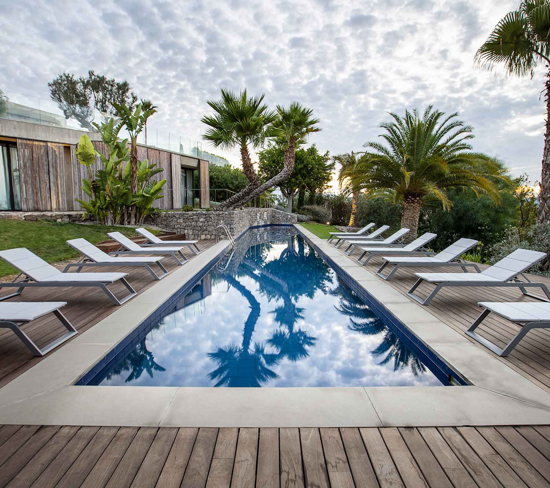 Scène sereine avec une piscine entourée de chaises longues et de palmiers, reflétant le ciel et la verdure luxuriante.