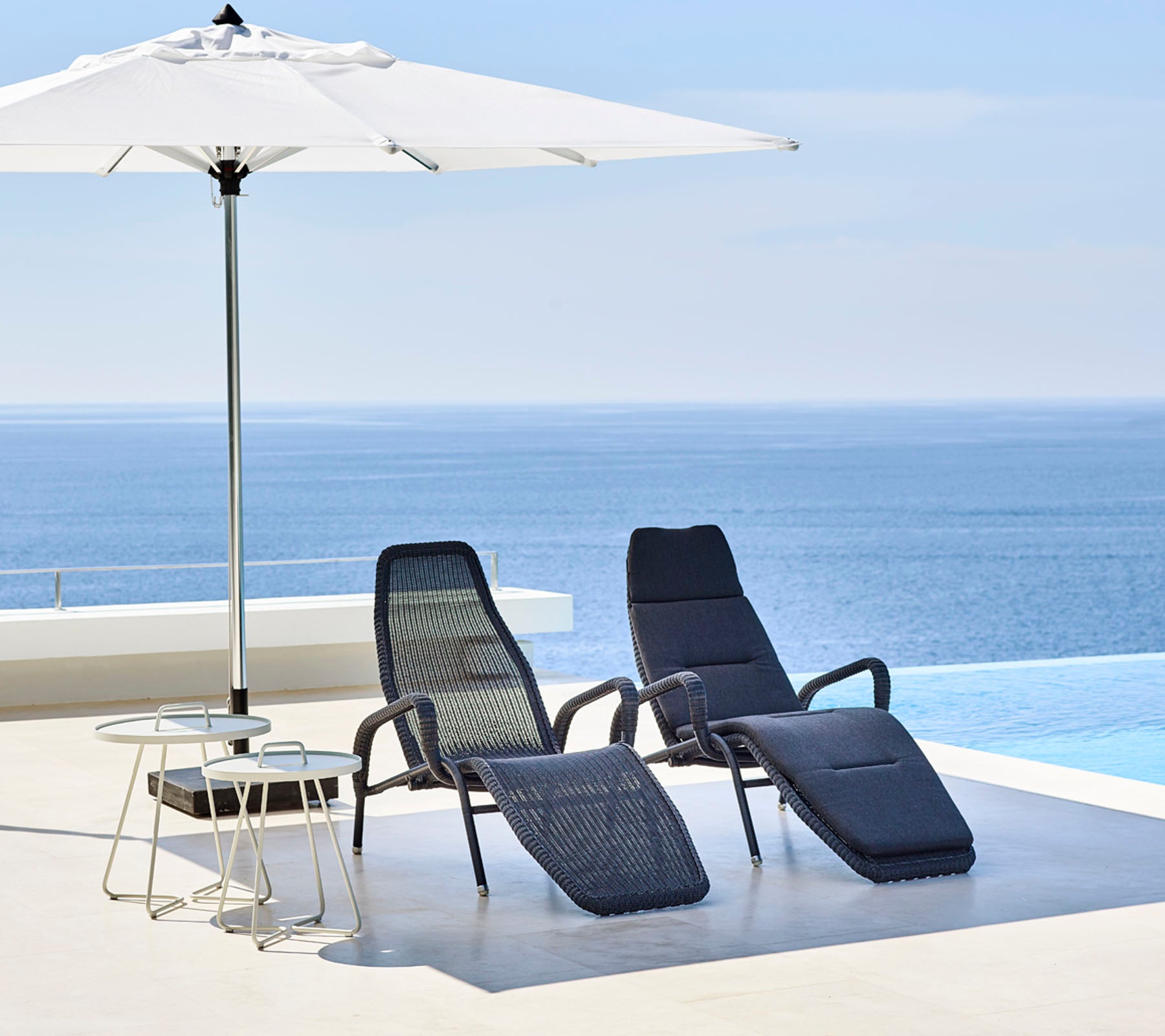 Deux chaises longues élégantes et une petite table se trouvent sous un grand parasol, avec une mer bleue sereine en arrière-plan.