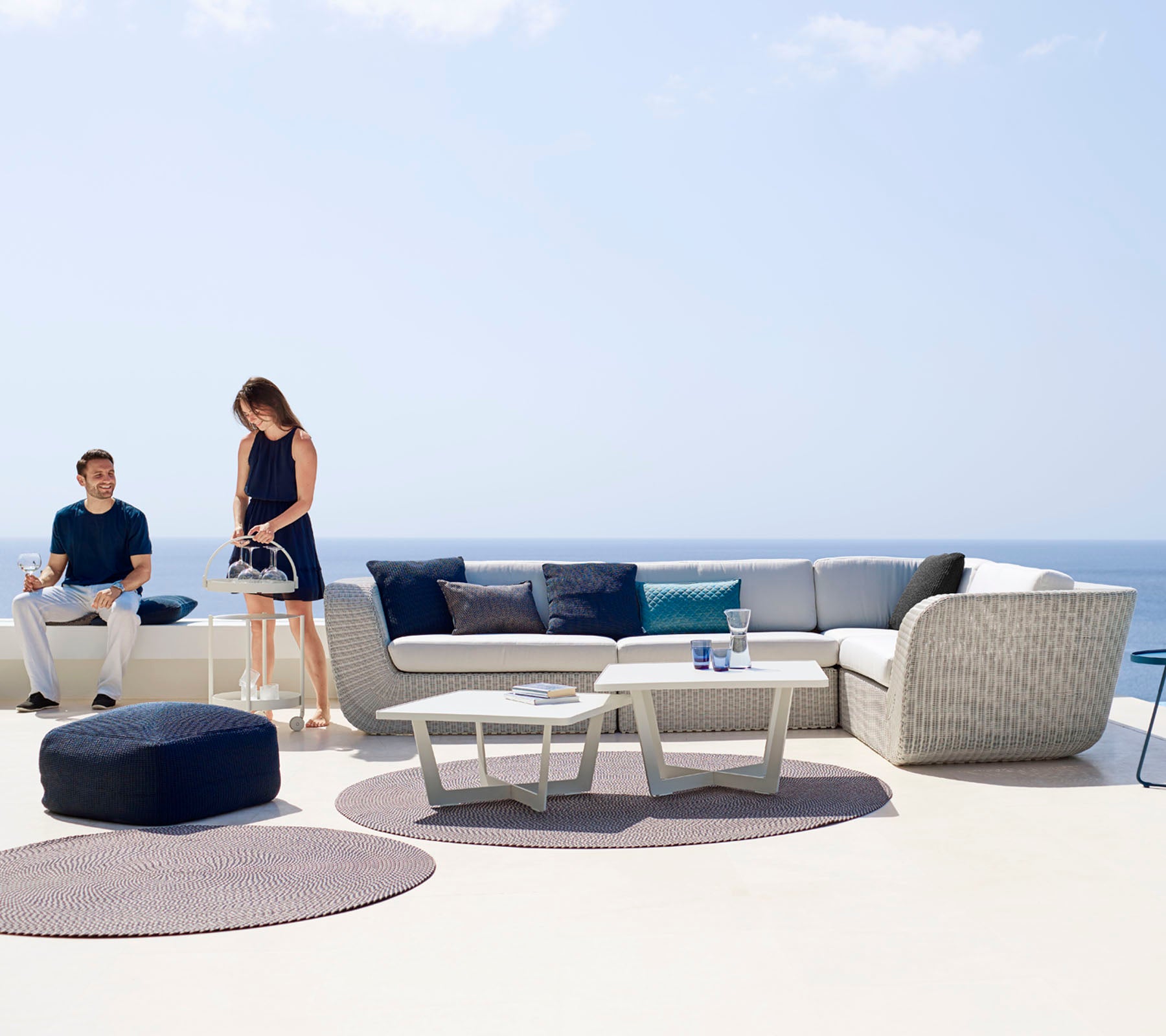 Un espace extérieur moderne avec un canapé d'angle, une table basse et des tapis décoratifs, avec deux personnes profitant de l'espace.