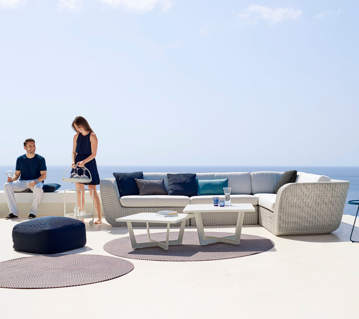 Un espace extérieur moderne avec un canapé d'angle, une table basse et des tapis décoratifs, avec deux personnes profitant de l'espace.